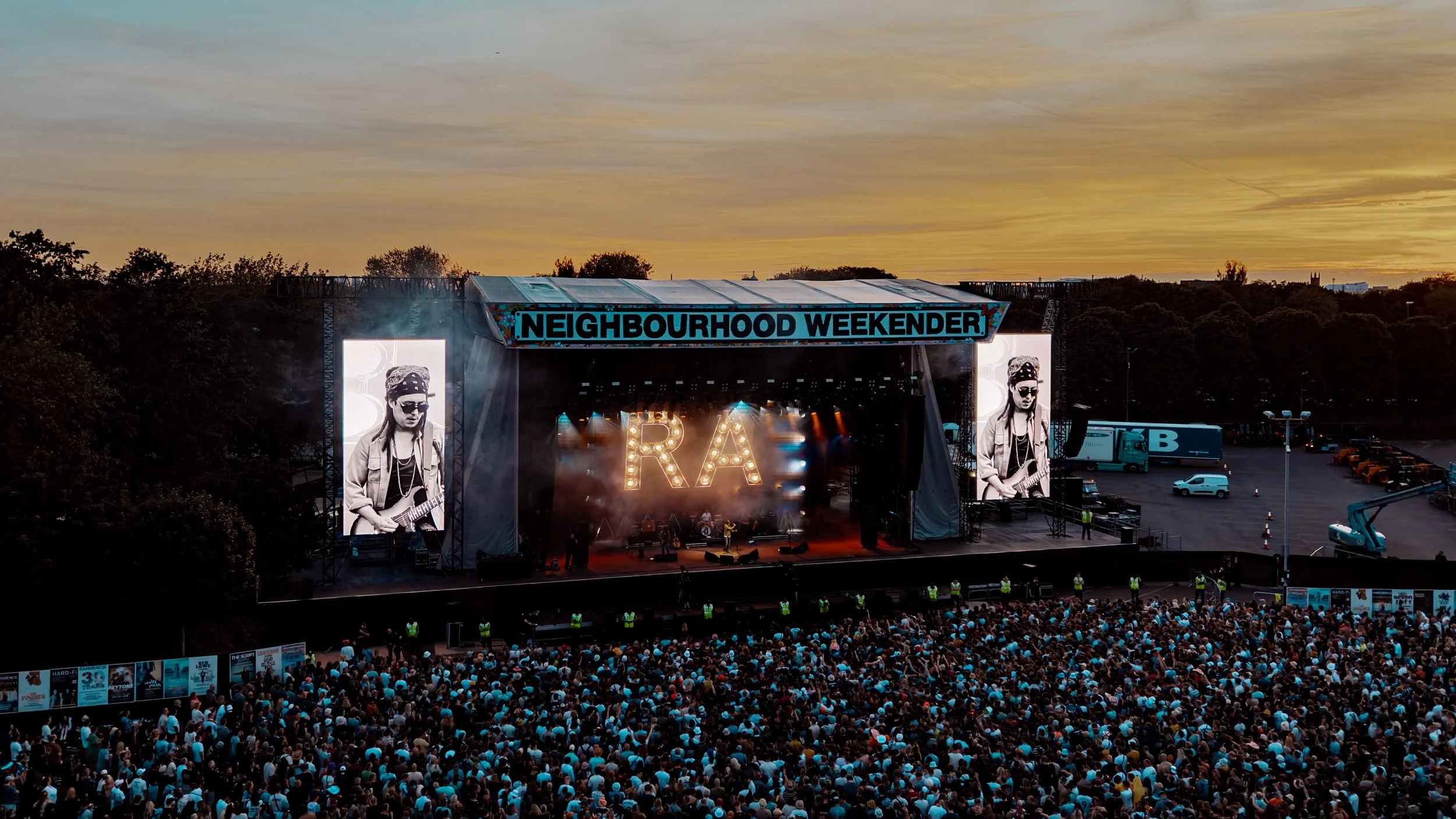 An outdoor concert stage at sunset with large screens showing musicians, crowded audience, and a banner reading Nighbourhood Weekender.