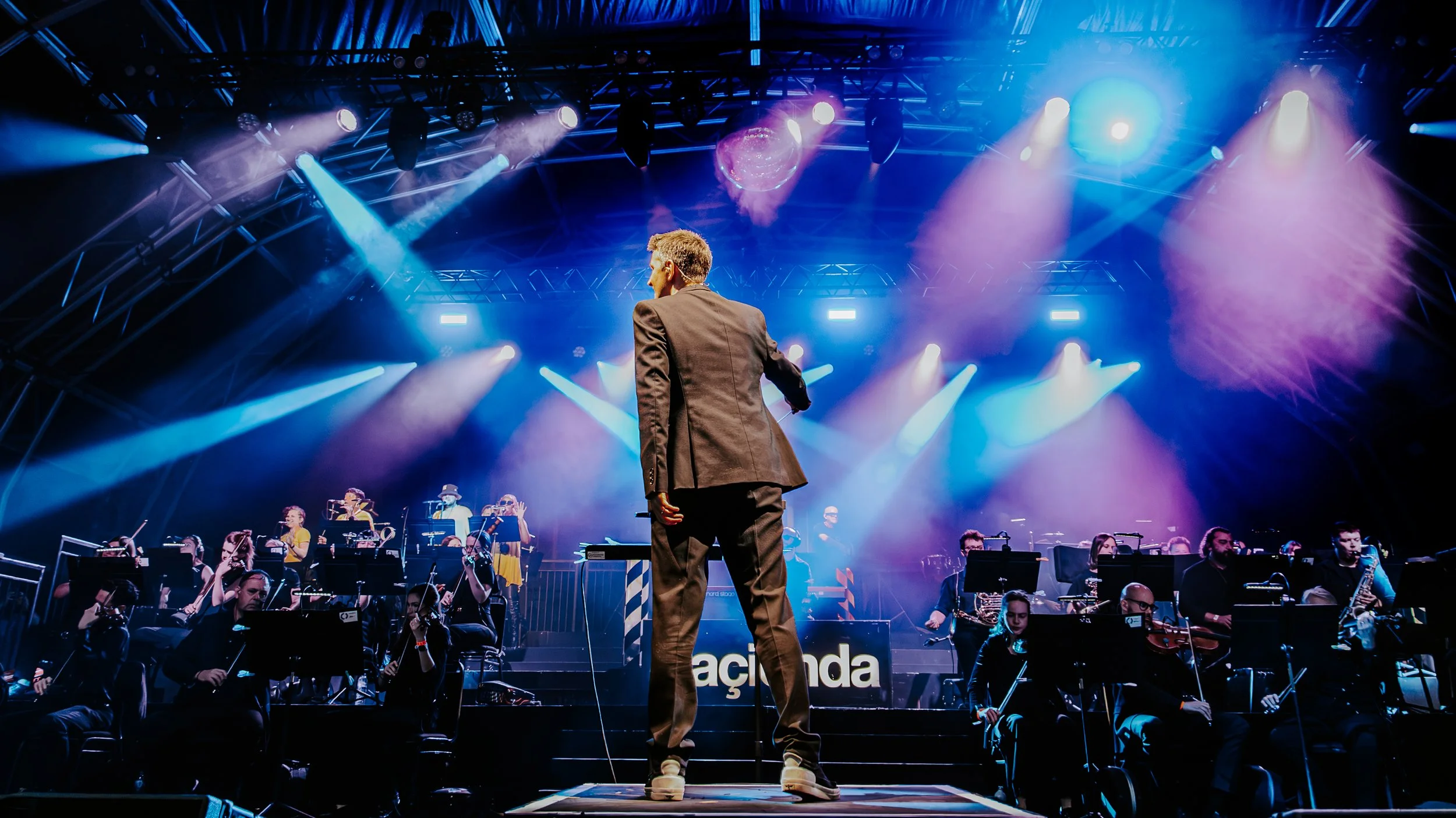 An orchestra performing on stage with colorful lights and a conductor in a suit facing the band members.