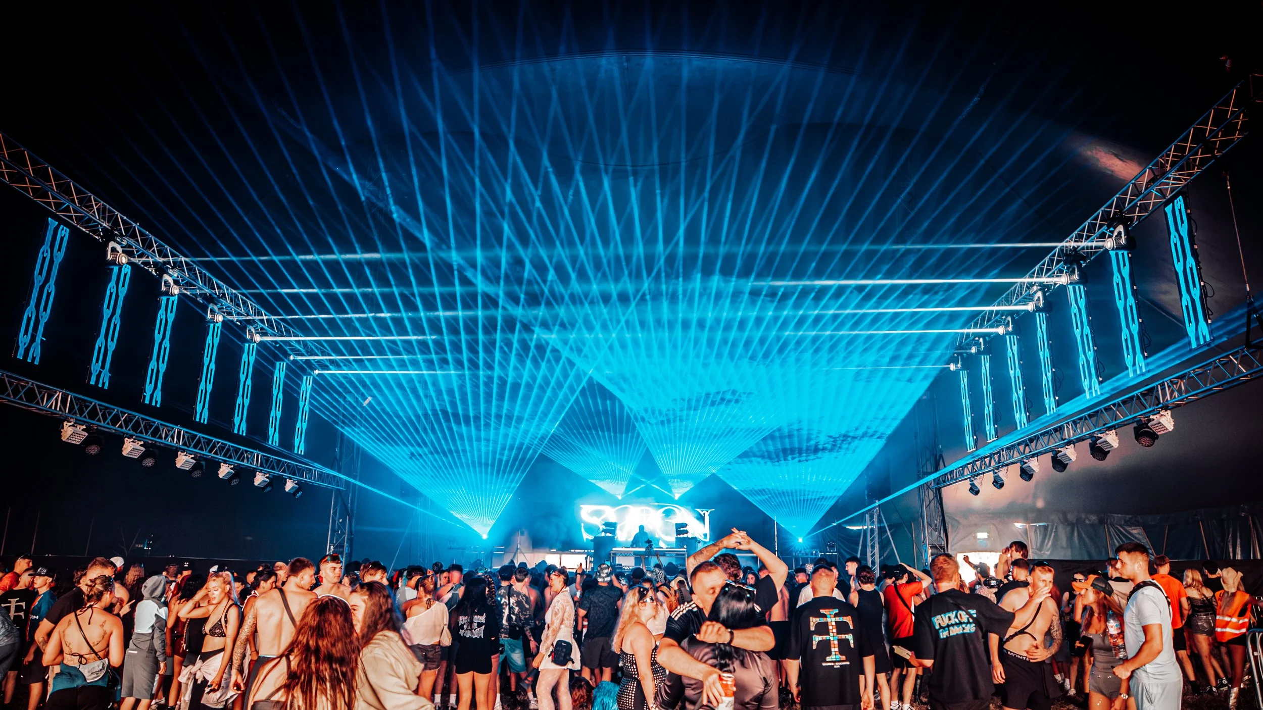 Crowd of people attending an outdoor music concert under a large blue laser light display with stage lighting and a DJ at the stage.