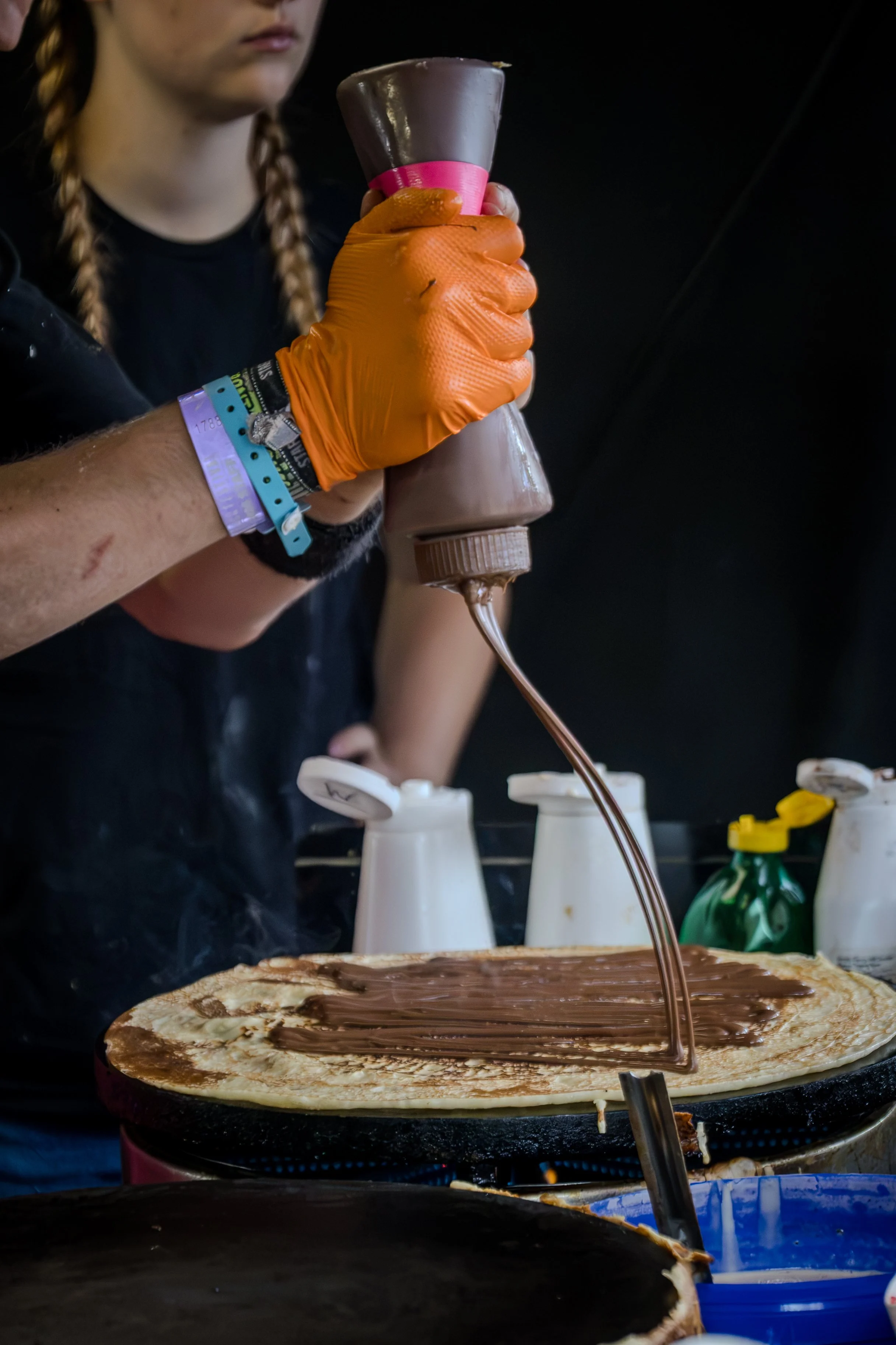 A person with braided hair and an orange glove is pouring chocolate topping over a large crepe on a griddle, with various condiment bottles in the background.