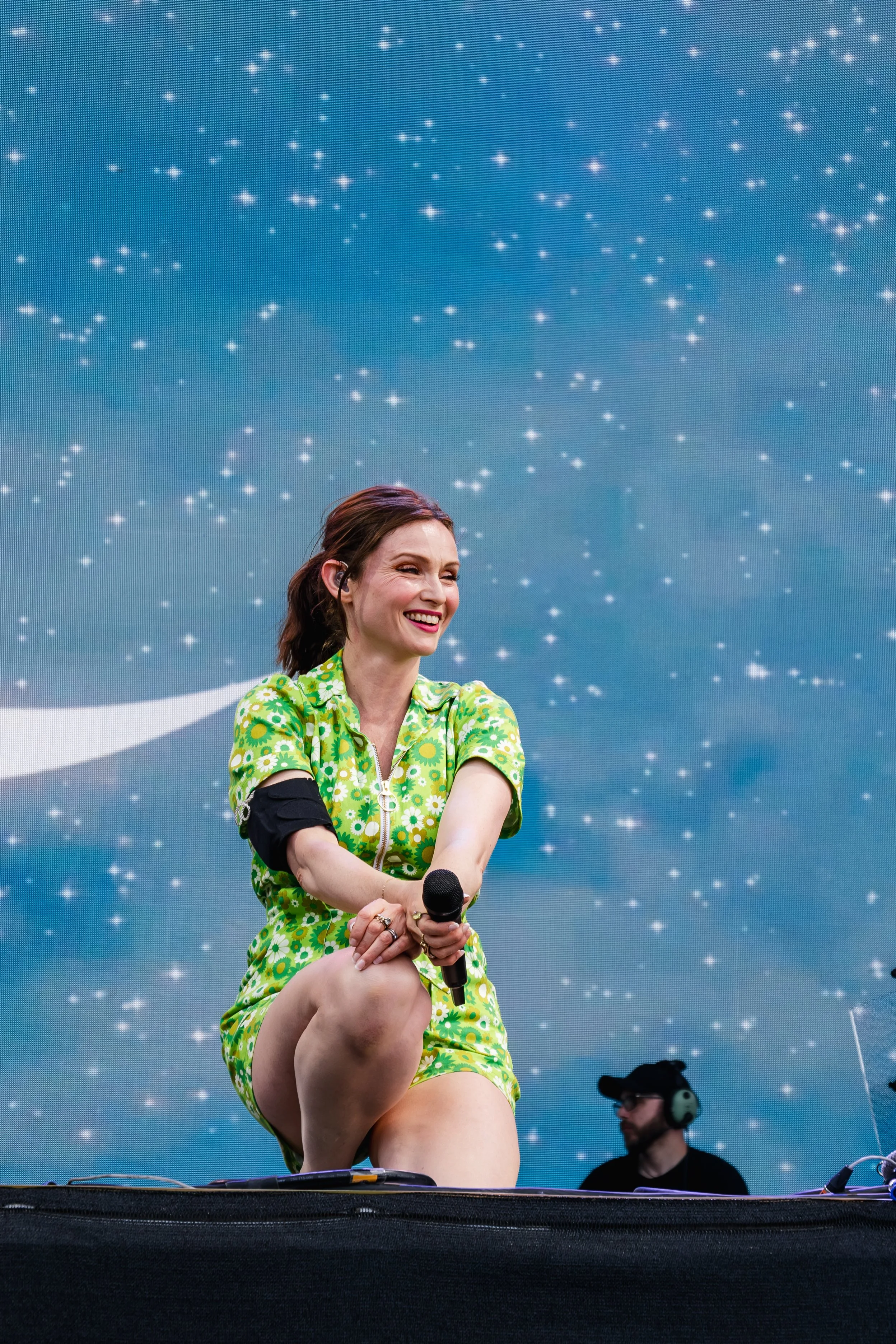 A woman in a bright green dress with flower patterns is on stage, kneeling with one knee up, holding a microphone and smiling. She has shoulder-length brown hair and is in front of a large LED screen displaying a starry night sky. A man with headphon