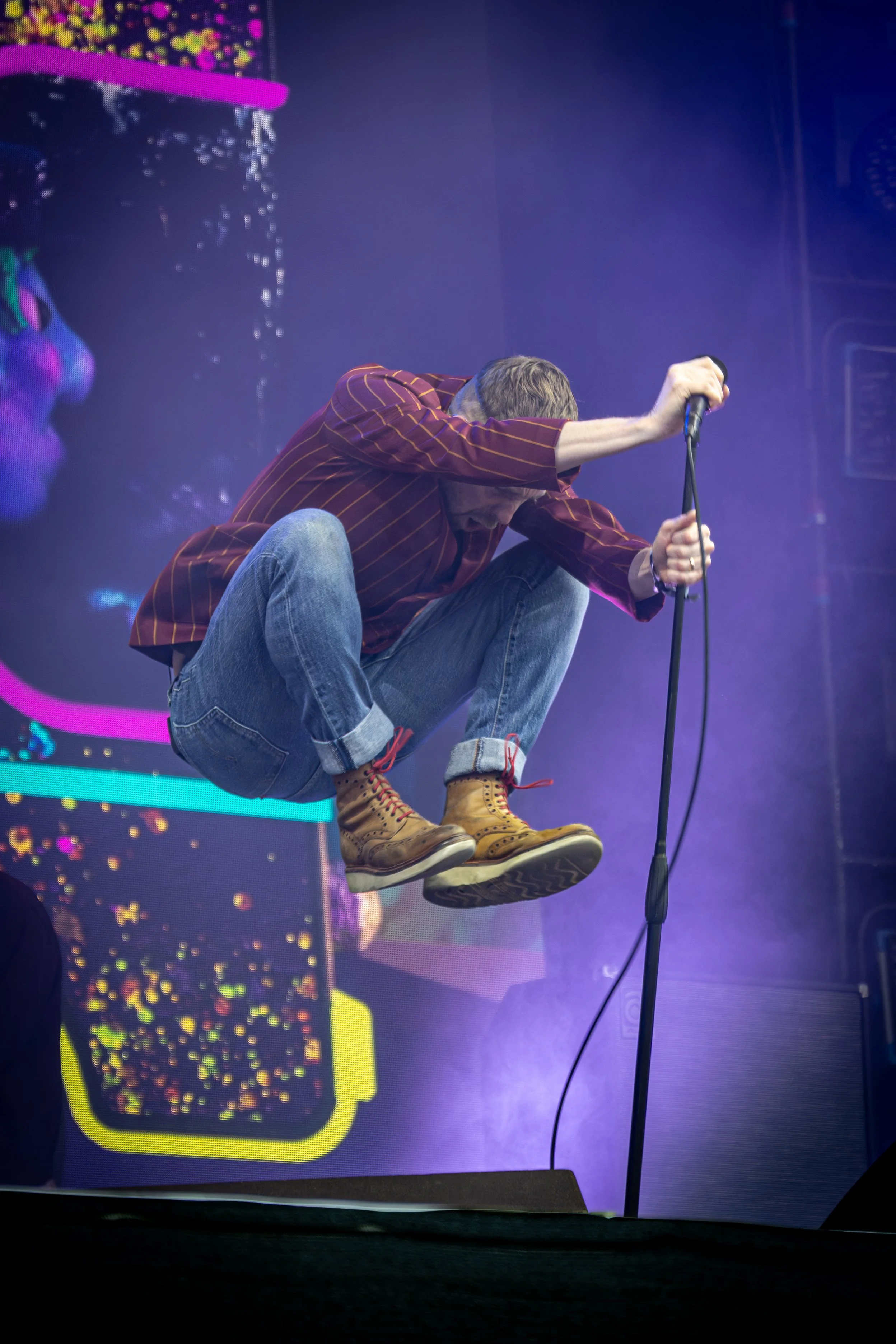 A man with gray hair and a beard is crouching and holding a microphone on stage. He is wearing a striped maroon shirt, blue jeans, and tan boots. The background features colorful, pixelated digital graphics and purple stage lighting.