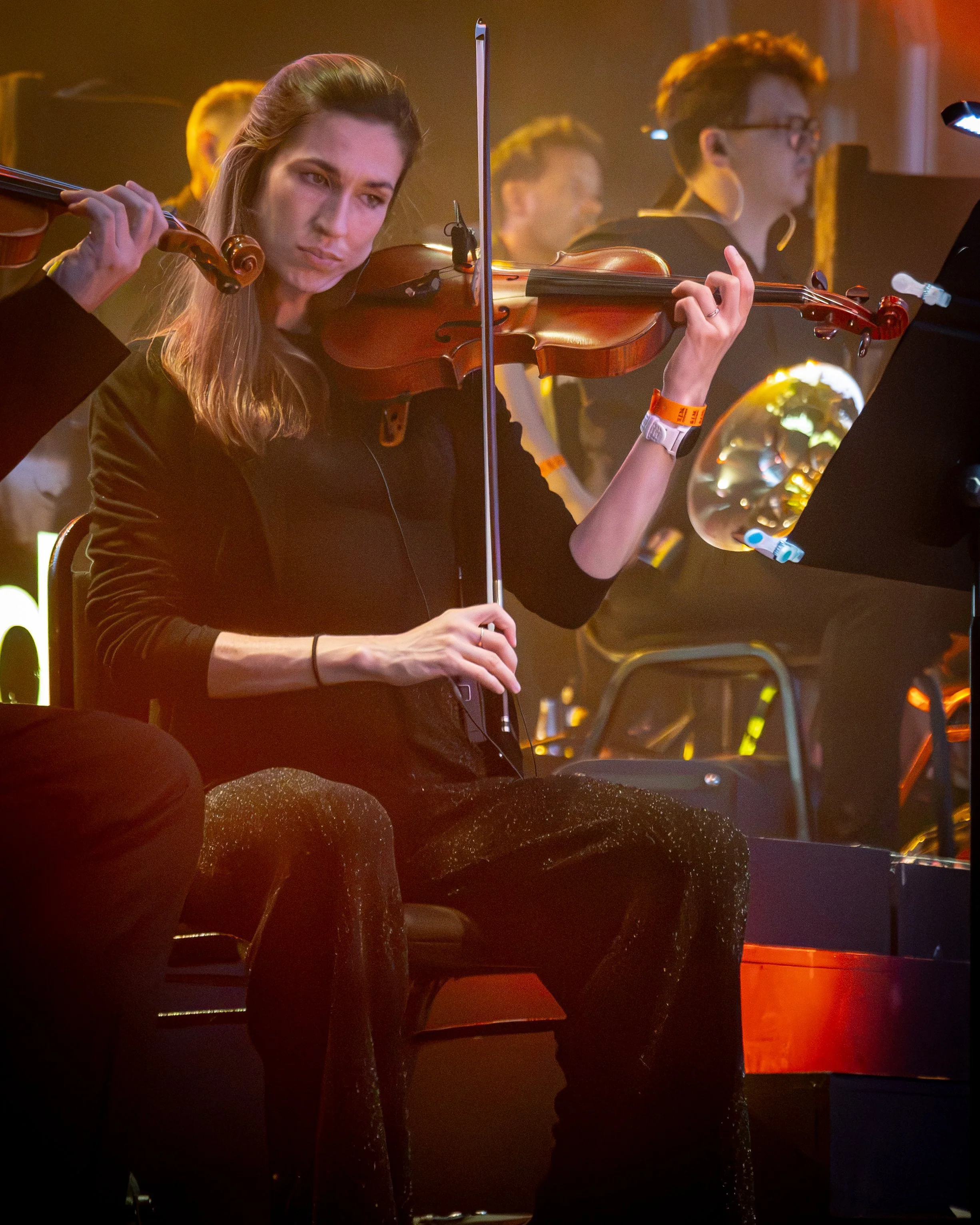 A woman playing the violin on stage, focused on her performance with a music stand in front of her. Several other musicians are visible in the background.