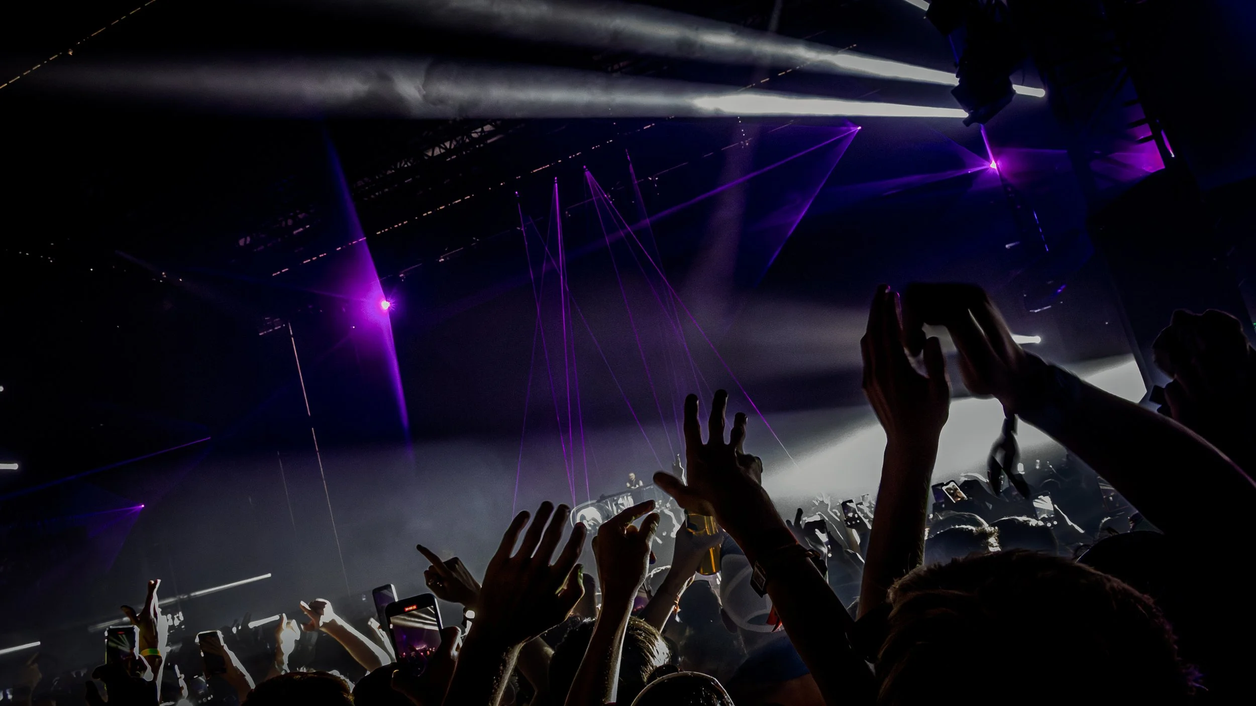 Crowd of concert attendees with hands raised, illuminated by purple and white laser lights in a dark indoor venue.