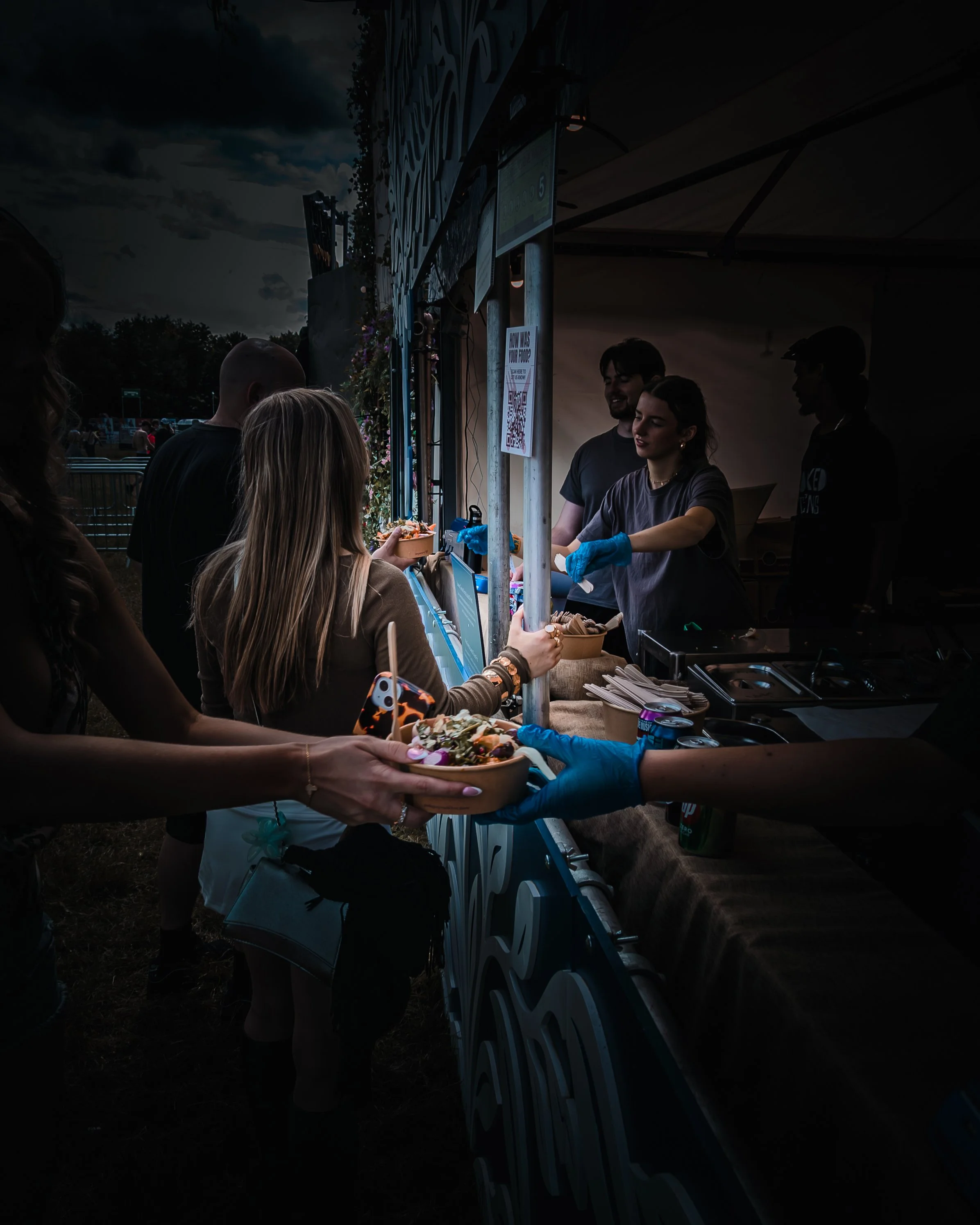 People at an outdoor food stall serving food during evening, with a dark cloudy sky overhead.
