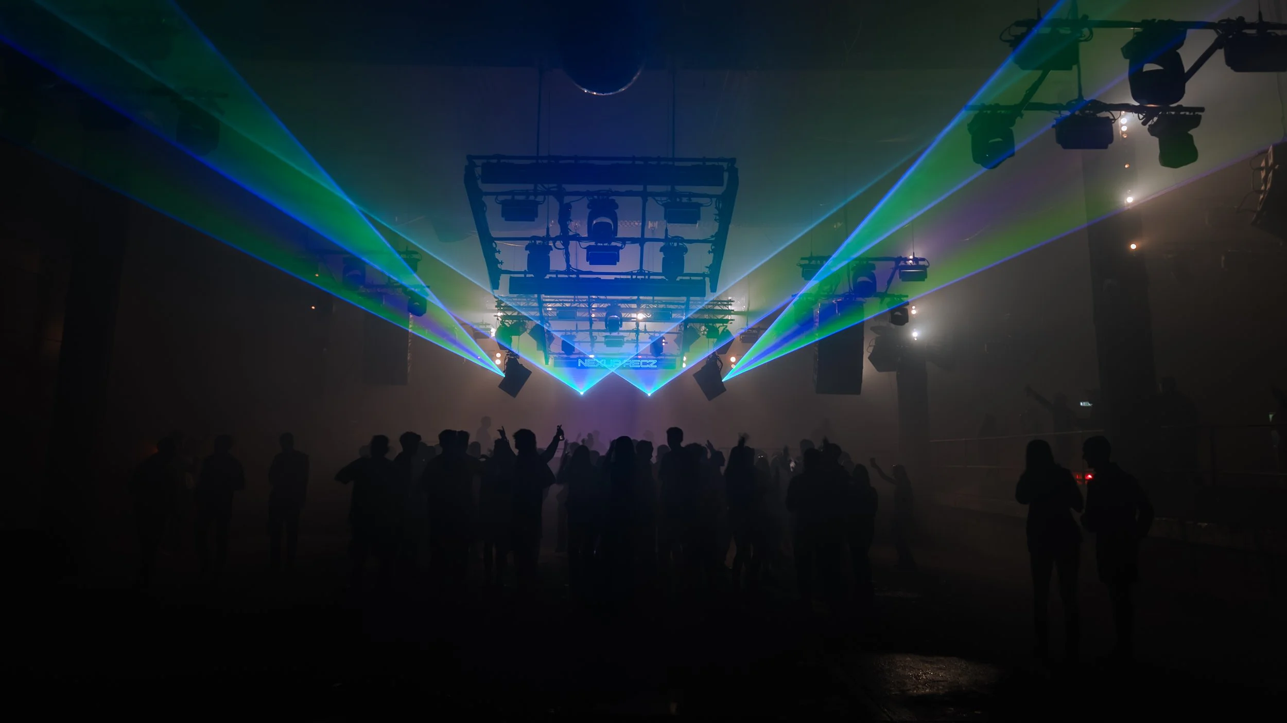 Concert or nightclub scene with colorful laser lights, silhouettes of people dancing and socializing, and stage equipment hanging from above.