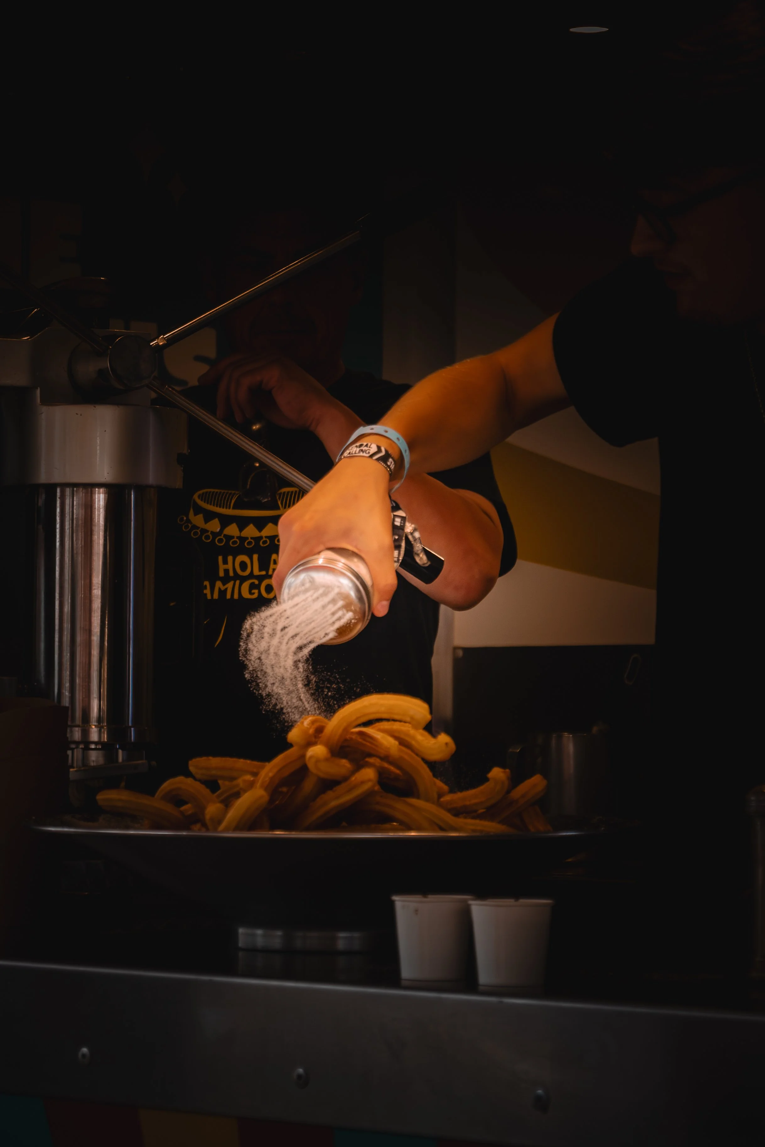 Person pouring powdered sugar onto a pile of cooked churros at a food stand.