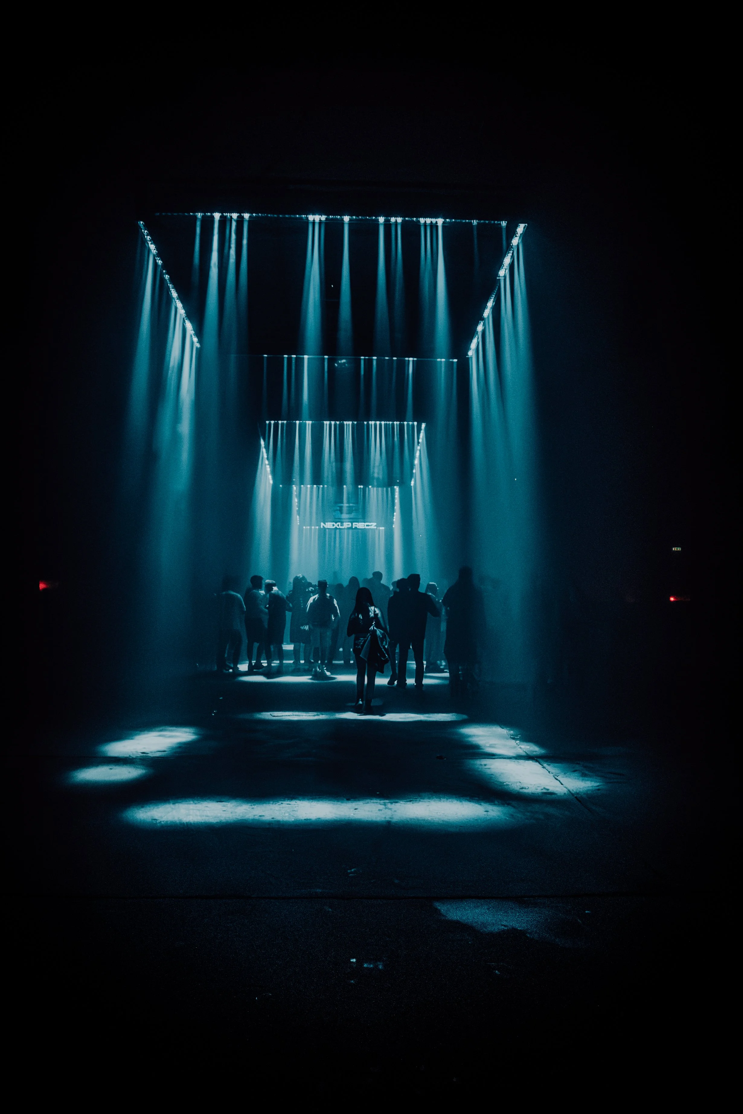 Dark scene with blue lighting and a group of people standing under a ceiling with hanging lights, with reflections on the wet floor.