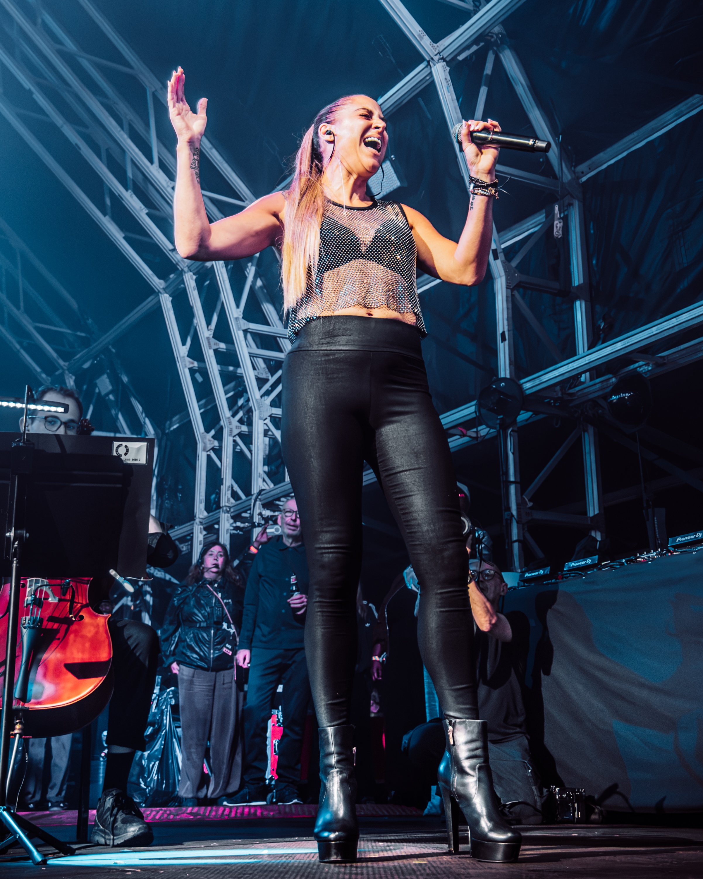 Female singer performing on stage with a microphone, wearing a black mesh top and leather pants, and large black platform boots, with musicians and audience members in the background.