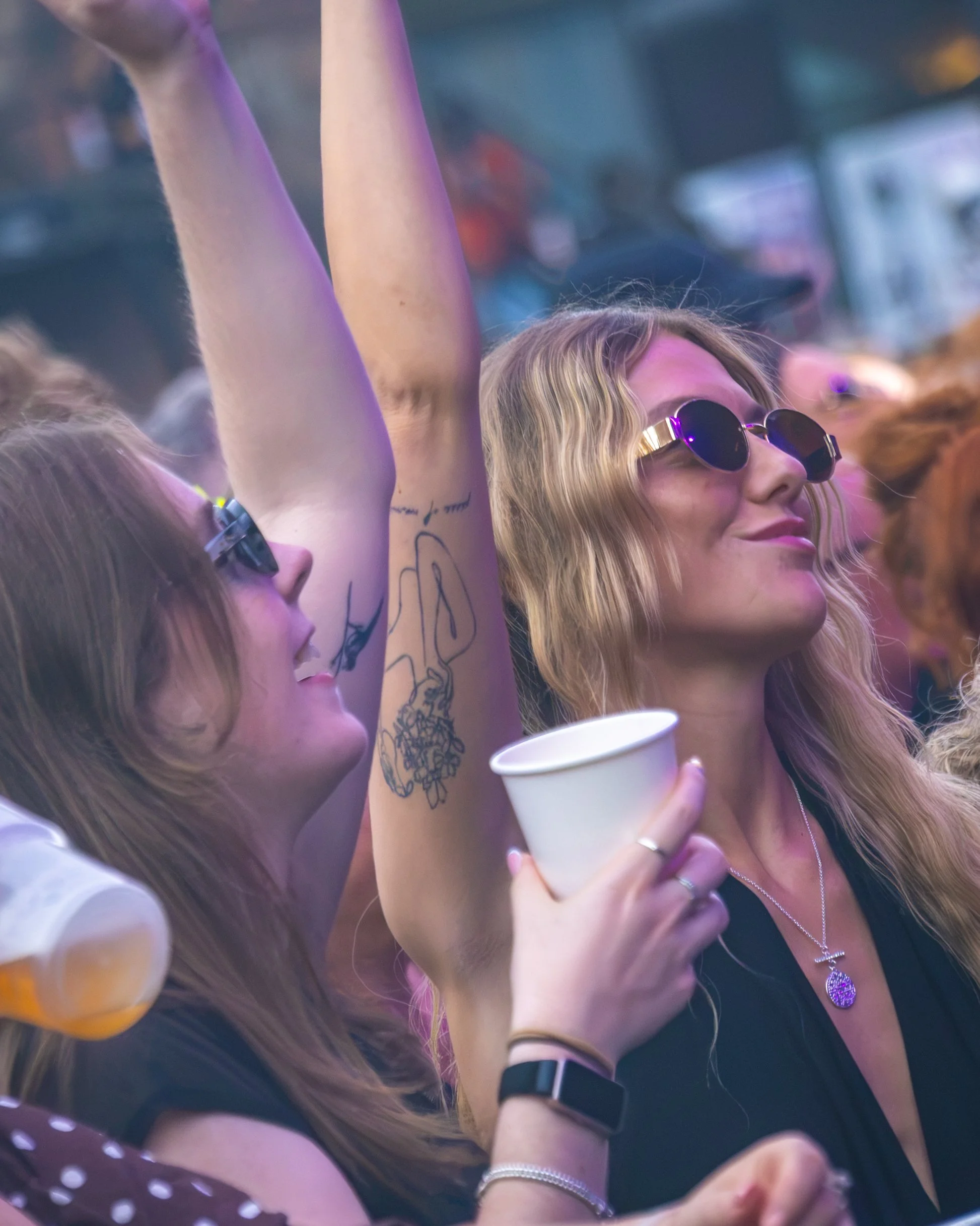 A woman with long blonde hair wearing sunglasses, a black top, and a necklace with a large round pendant, holding a white paper cup, at a lively event with others around her.