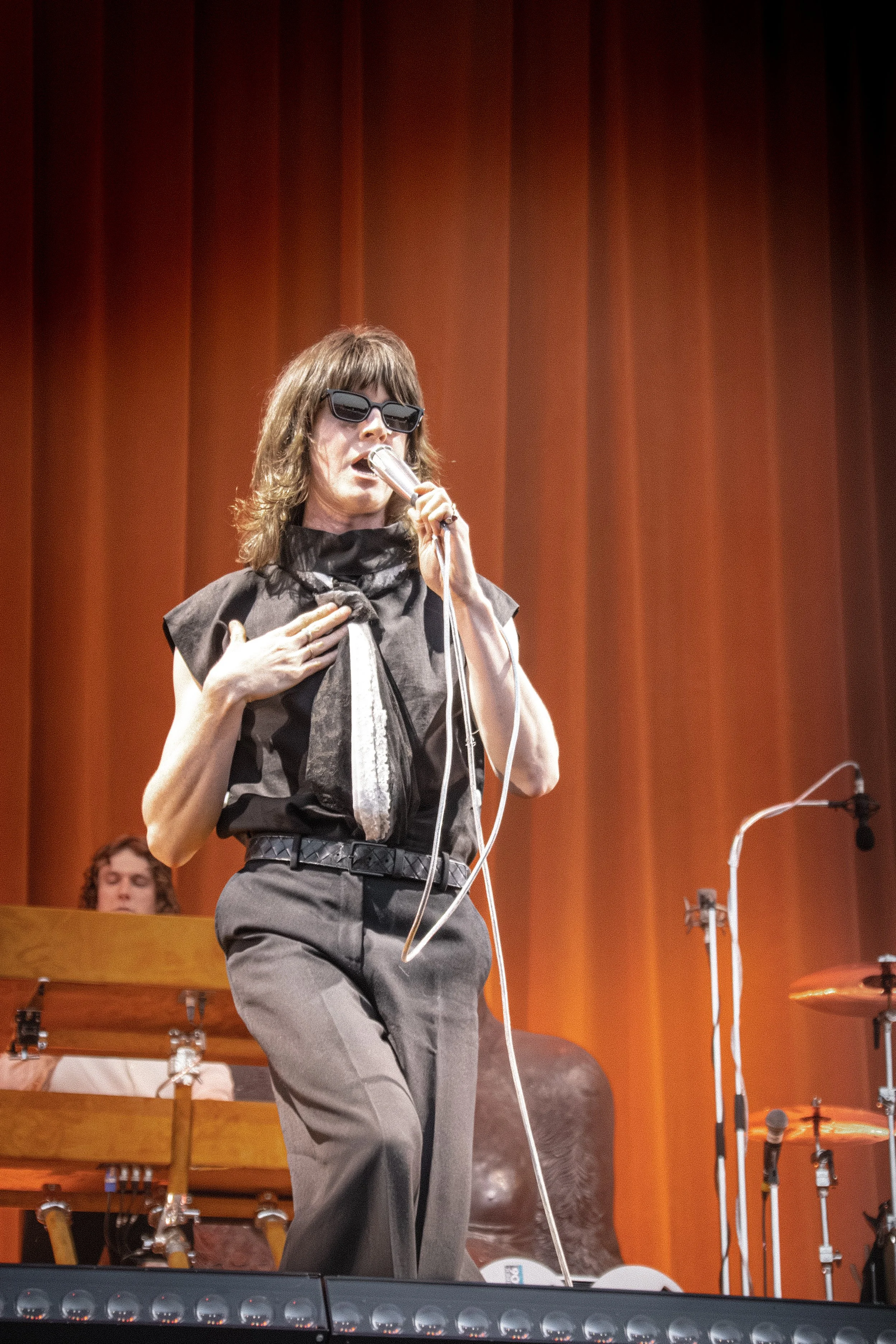 A woman with wavy medium-length hair, wearing dark sunglasses, a black top with a patterned scarf tied around her neck, and grey high-waisted pants, holding a microphone and singing on stage against a backdrop of orange curtains. There are music stan