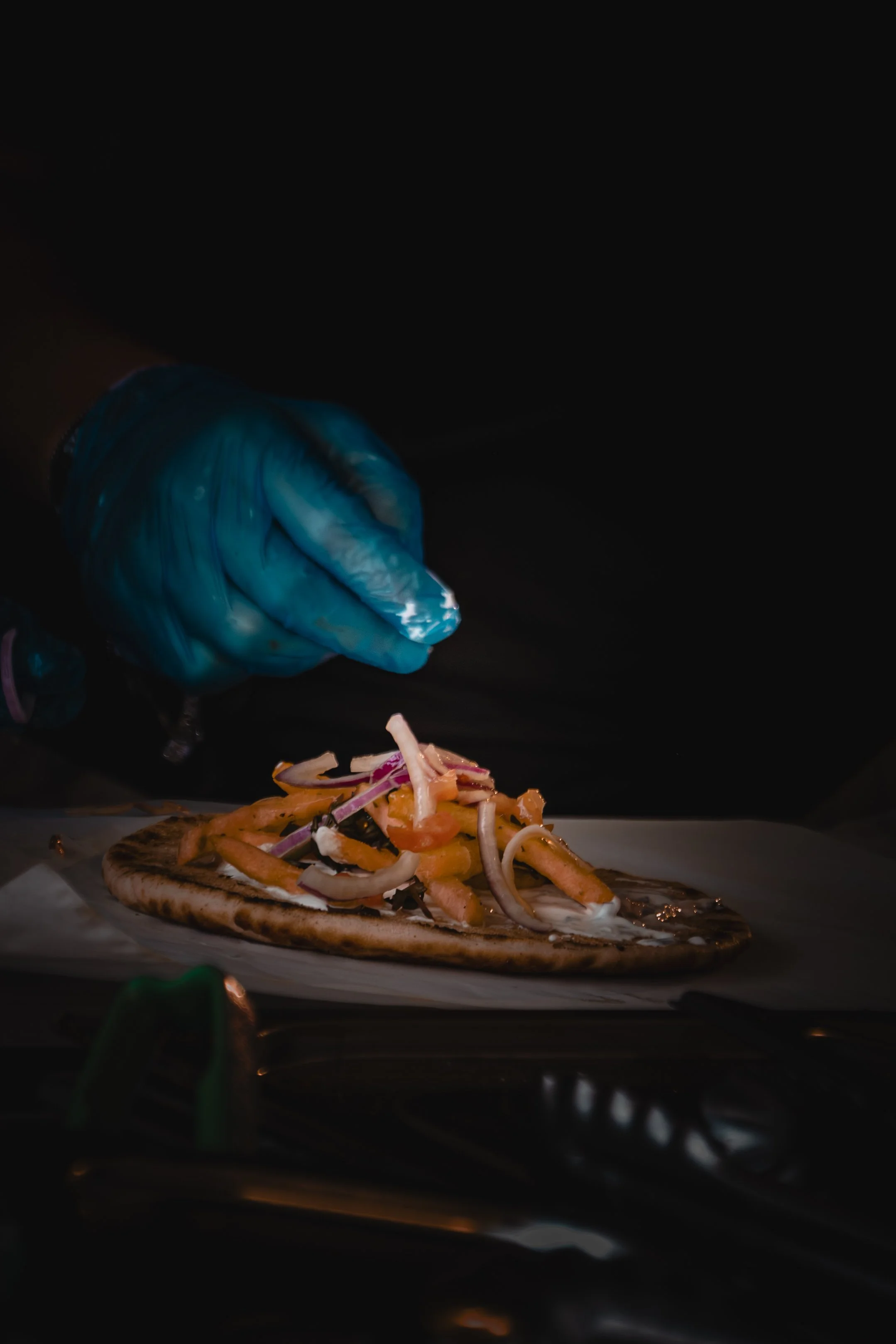 A person wearing a blue disposable glove is preparing to place toppings on a pizza, which sits on a baking sheet lined with parchment paper. The pizza has cheese and various vegetables such as onions and peppers. The background is dark, possibly a ki