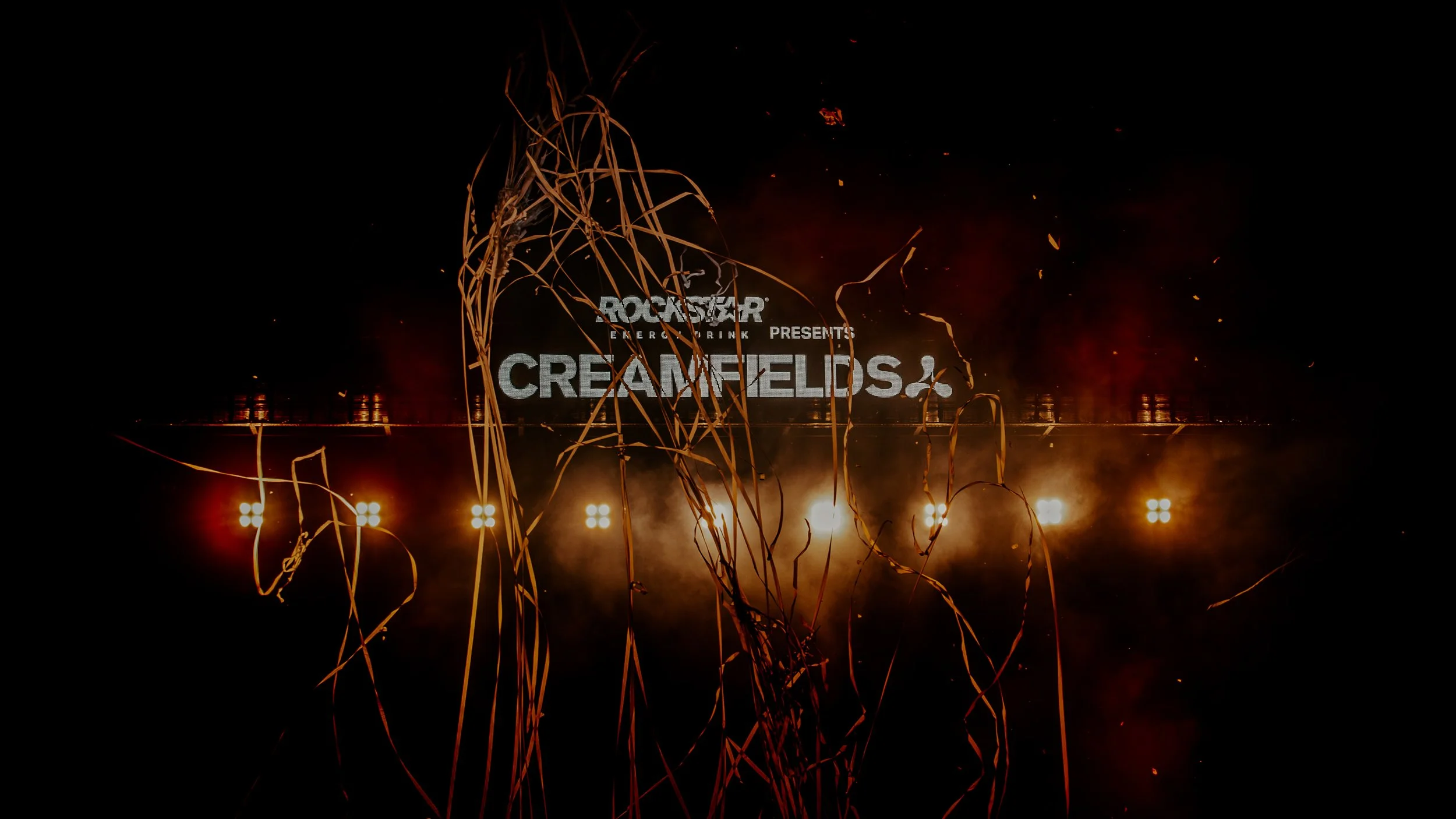 Concert stage with large screen displaying 'Rockstar Energy Drink presents Creamfields', surrounded by orange streamers and bright orange and white lights.