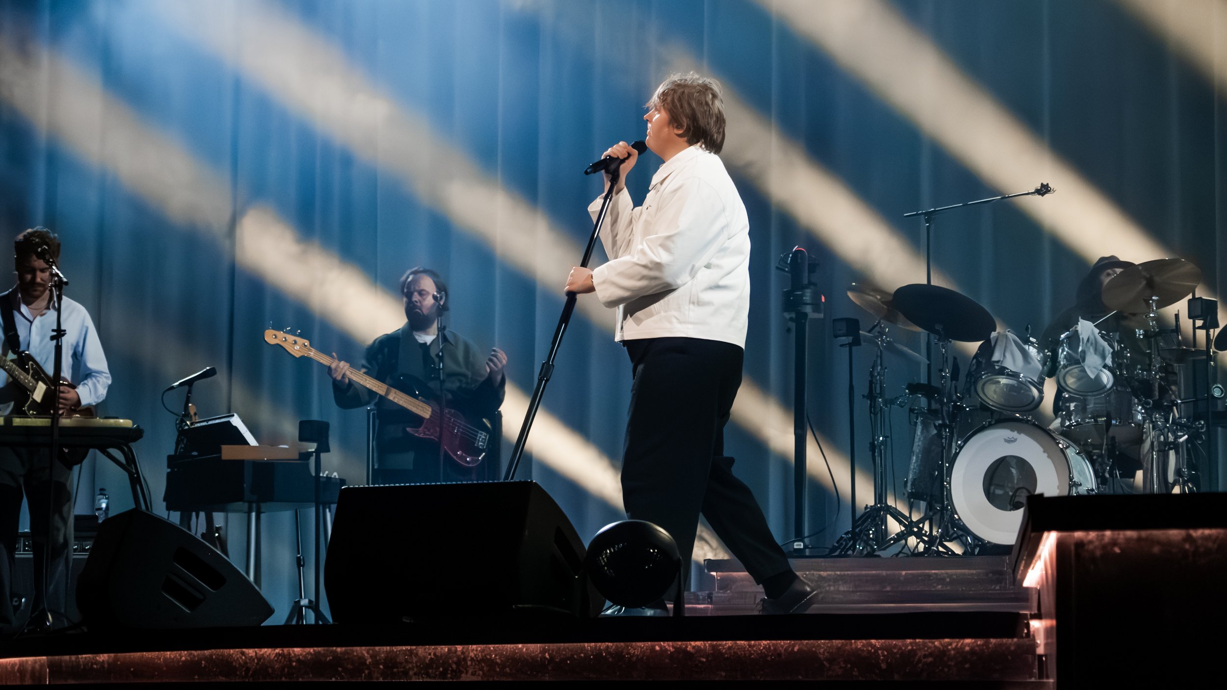 A person singing into a microphone on stage with a band playing instruments behind them, including a guitarist, bassist, keyboard, and drums, with stage lighting casting beams of light.
