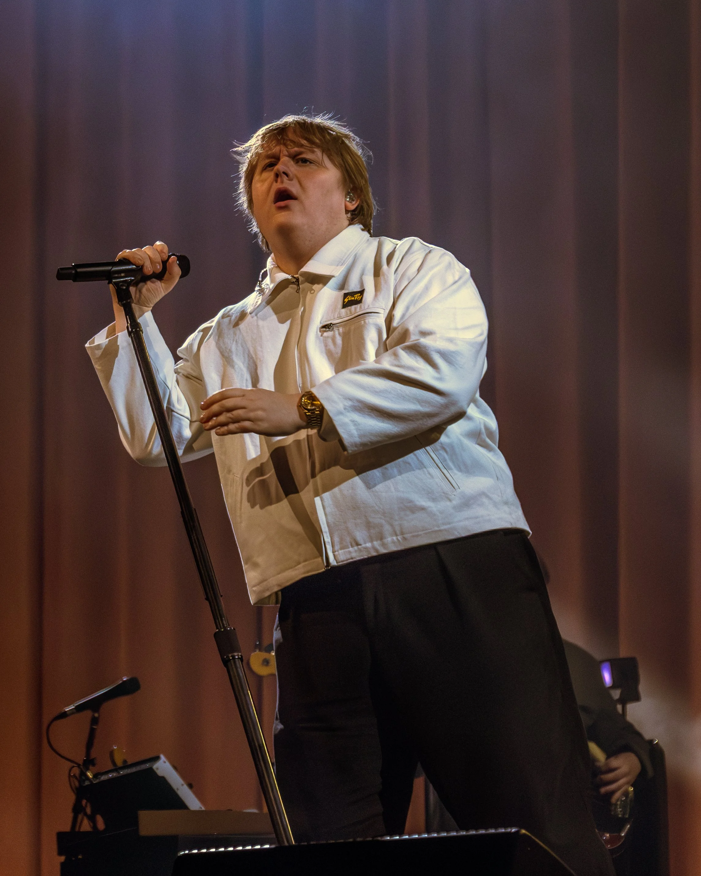 A man singing or speaking into a microphone on a stage with a dark curtain background, wearing a white jacket, a gold watch, and holding a glass in his right hand, while standing behind a keyboard.