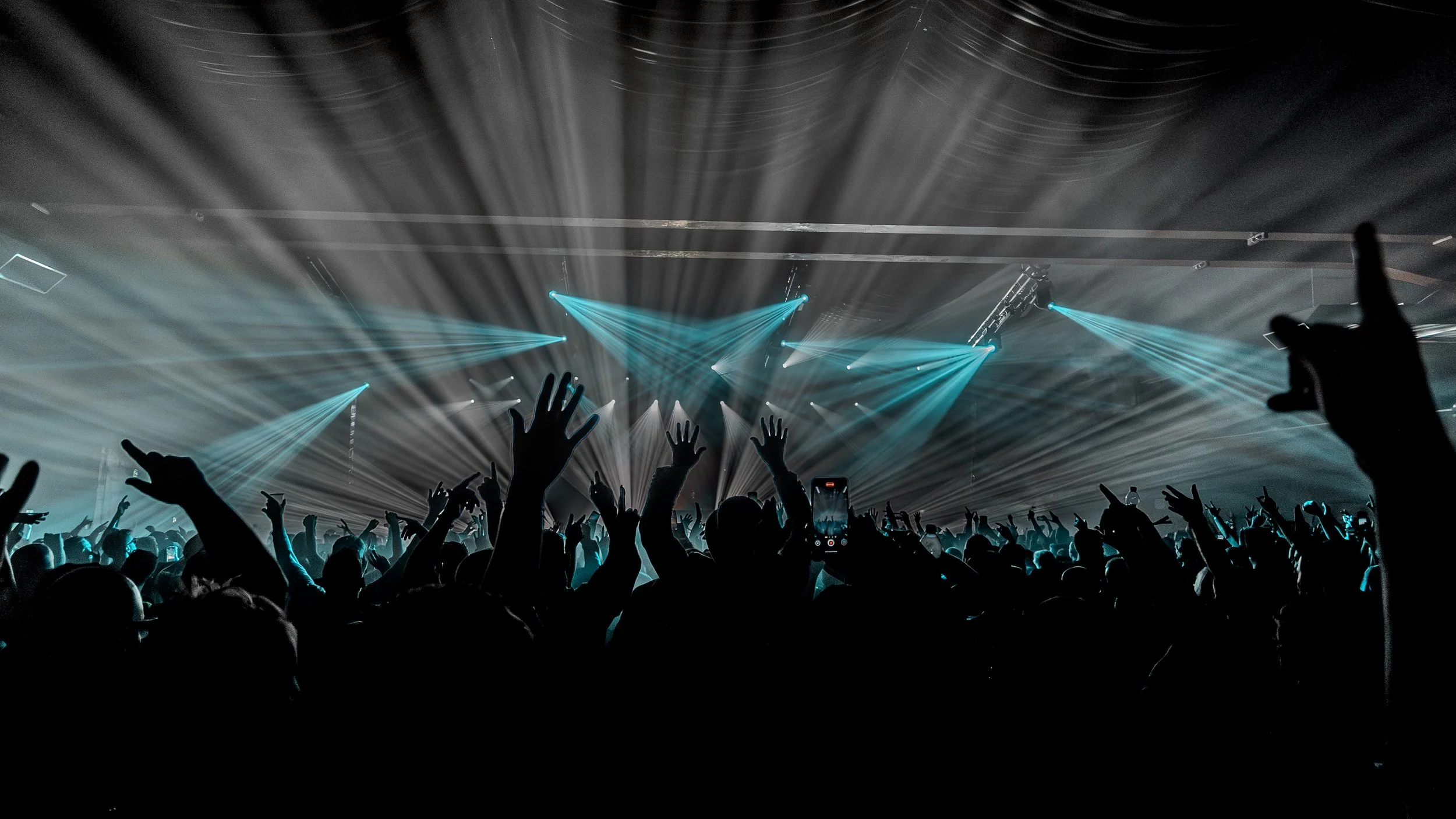 Crowd of people at a concert or dance event with hands raised and blue light beams shining from the stage.