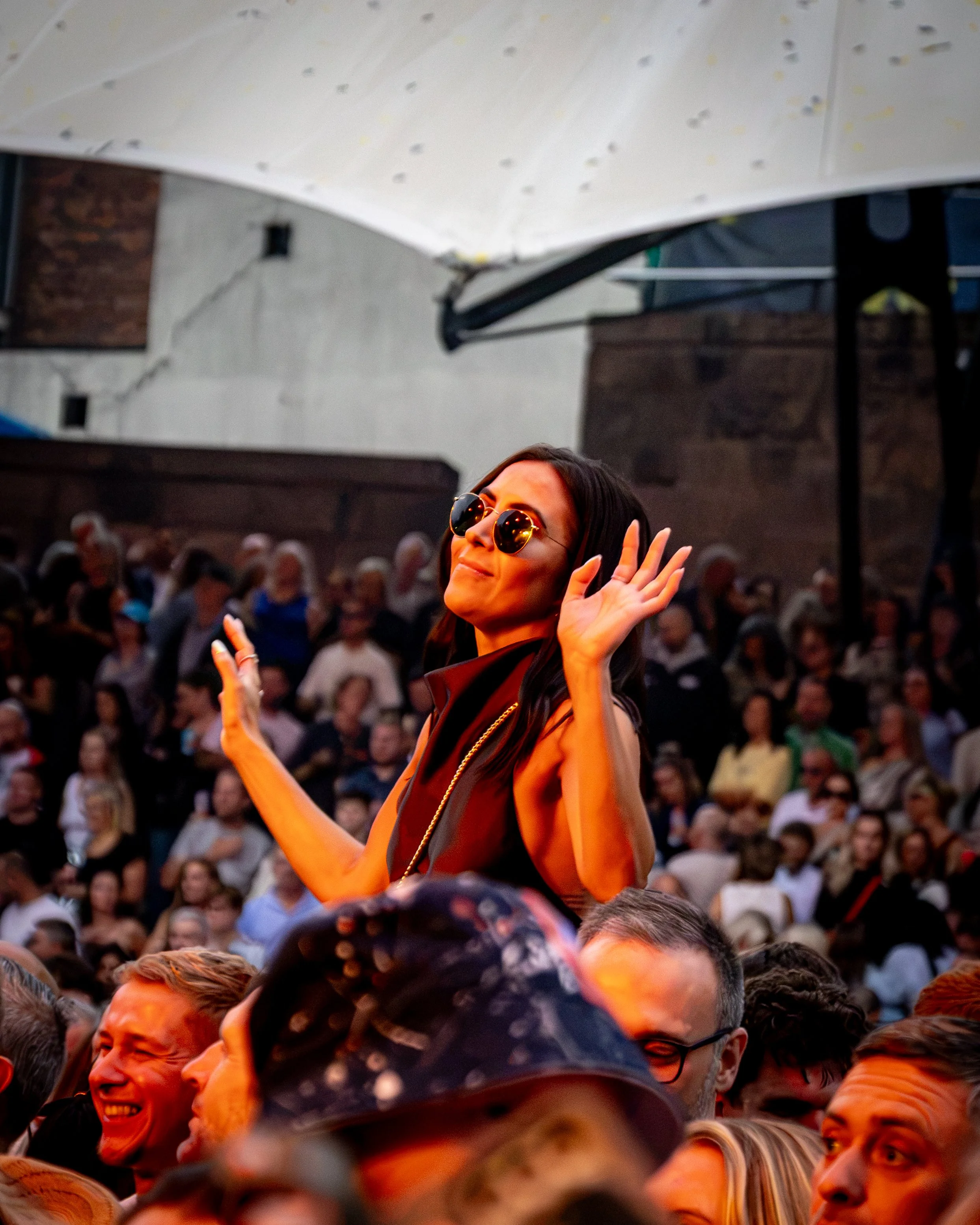 A woman with dark hair wearing sunglasses and a sleeveless top, smiling and raising her hand, surrounded by a crowd of people at an outdoor event.