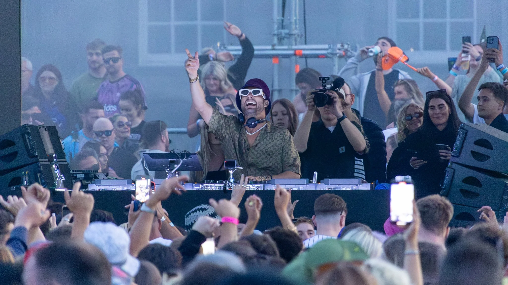 A DJ wearing white sunglasses and a patterned shirt is cheering with his arms raised at an outdoor music festival, surrounded by a crowd of people taking photos and videos.