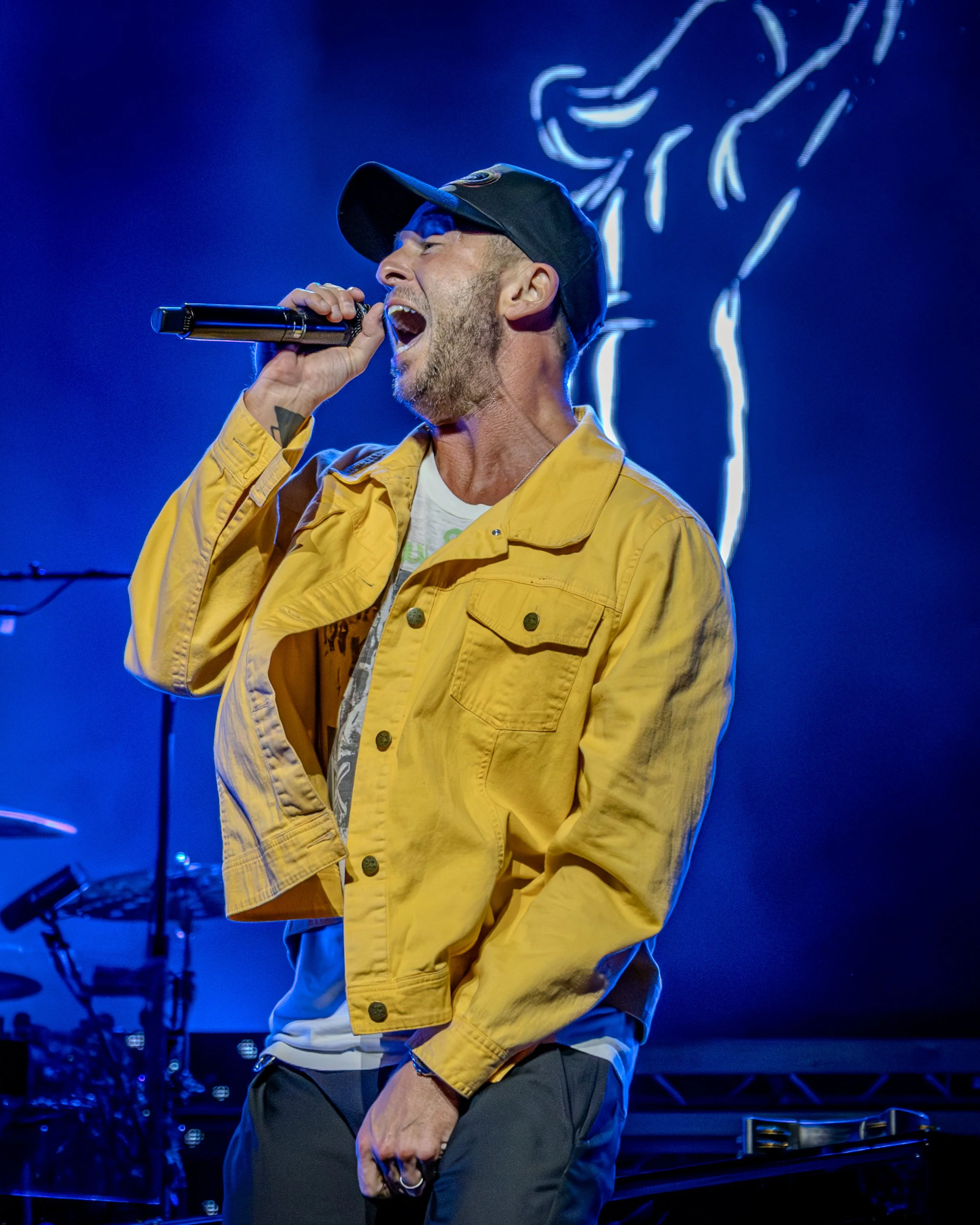 A man singing into a microphone on stage, wearing a baseball cap, yellow jacket, and dark pants, with a neon-like drawing of a microphone and hand in the background.