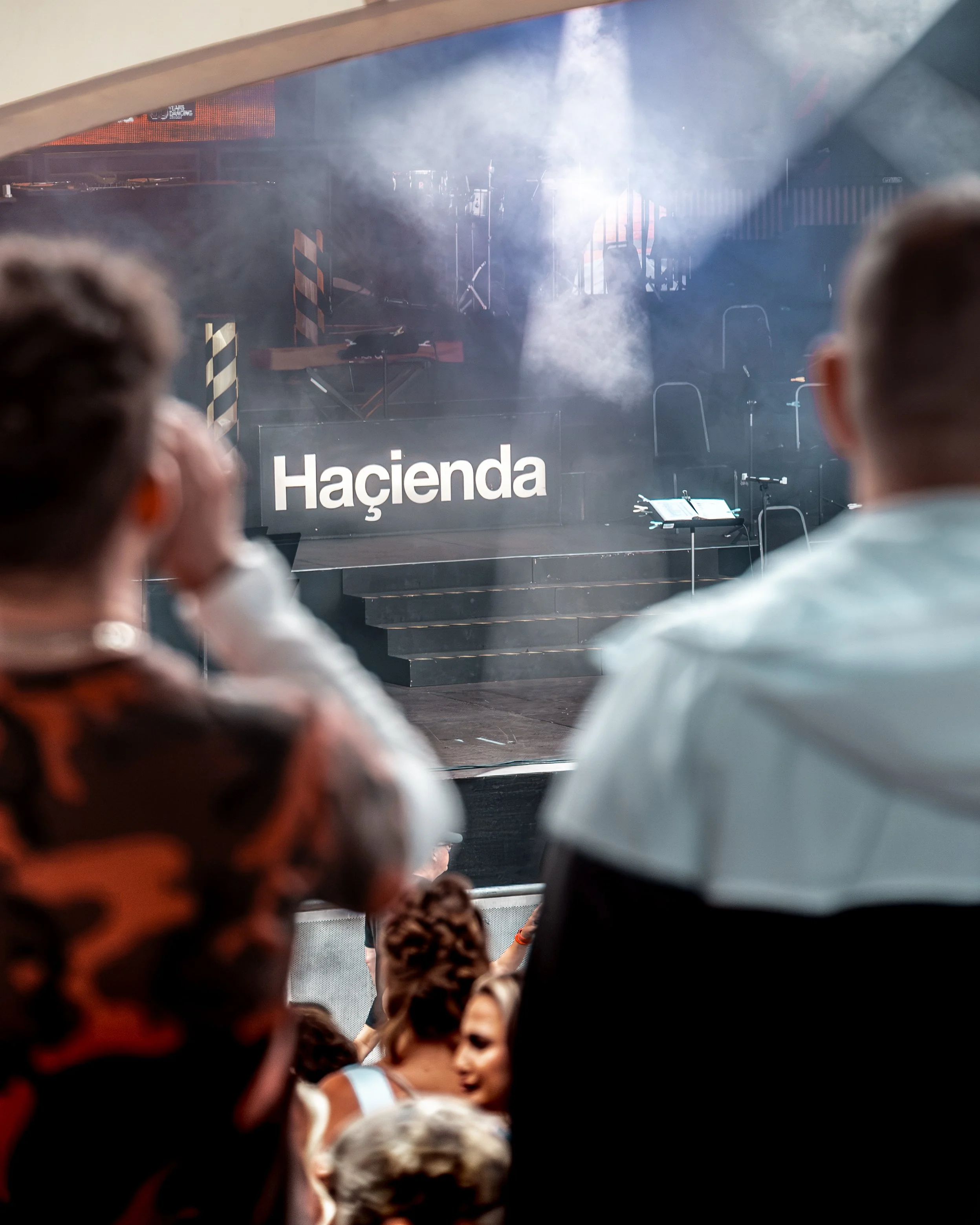 People attending an event or concert with stage lighting and equipment visible, framing the scene through an opening.