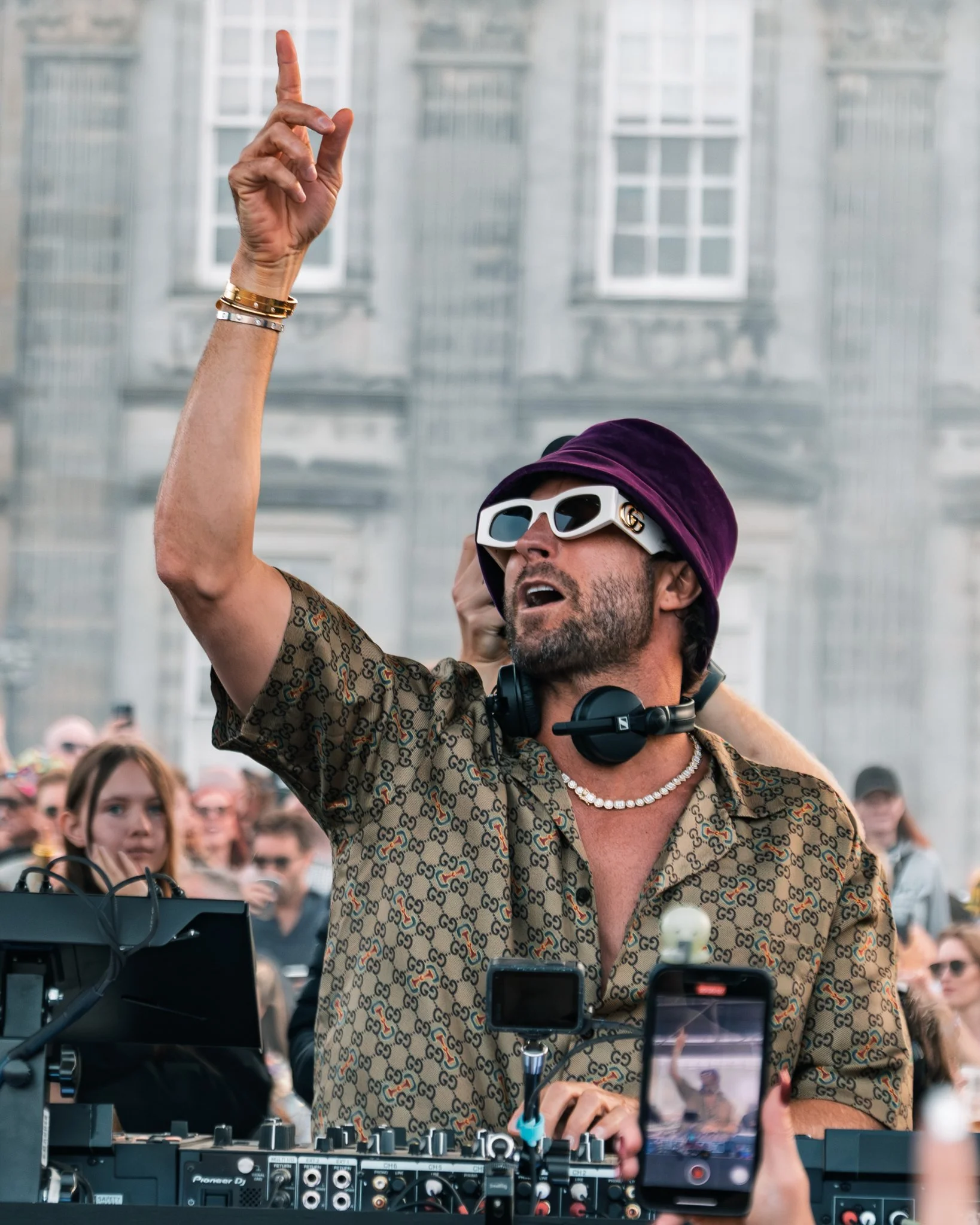 A DJ wearing a purple hat, patterned shirt, and sunglasses, standing behind DJ equipment at an outdoor event, raising one hand with a finger pointed upwards, crowd in the background.