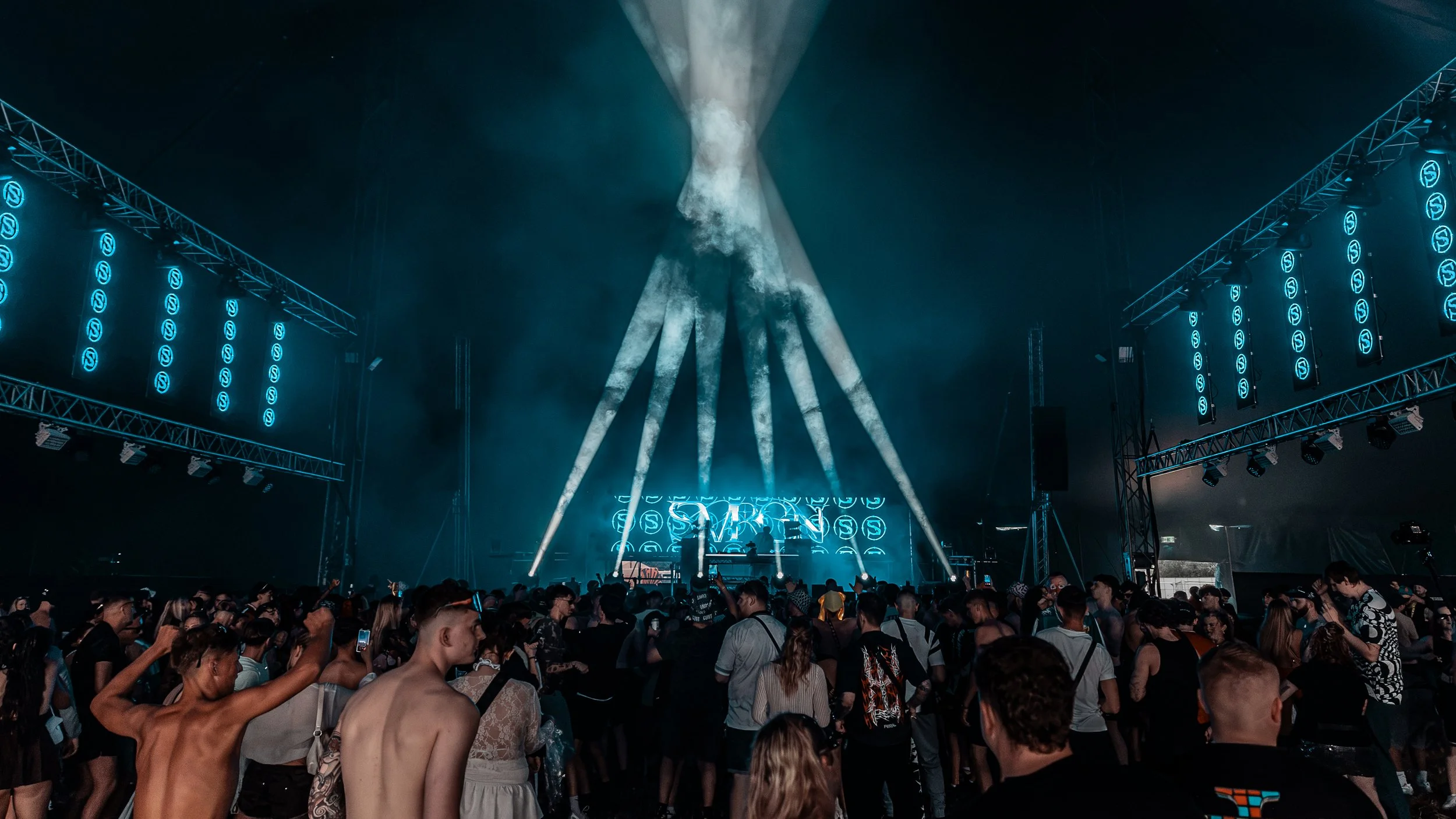 Crowd of people at a concert or music festival with stage lights and smoke effects.