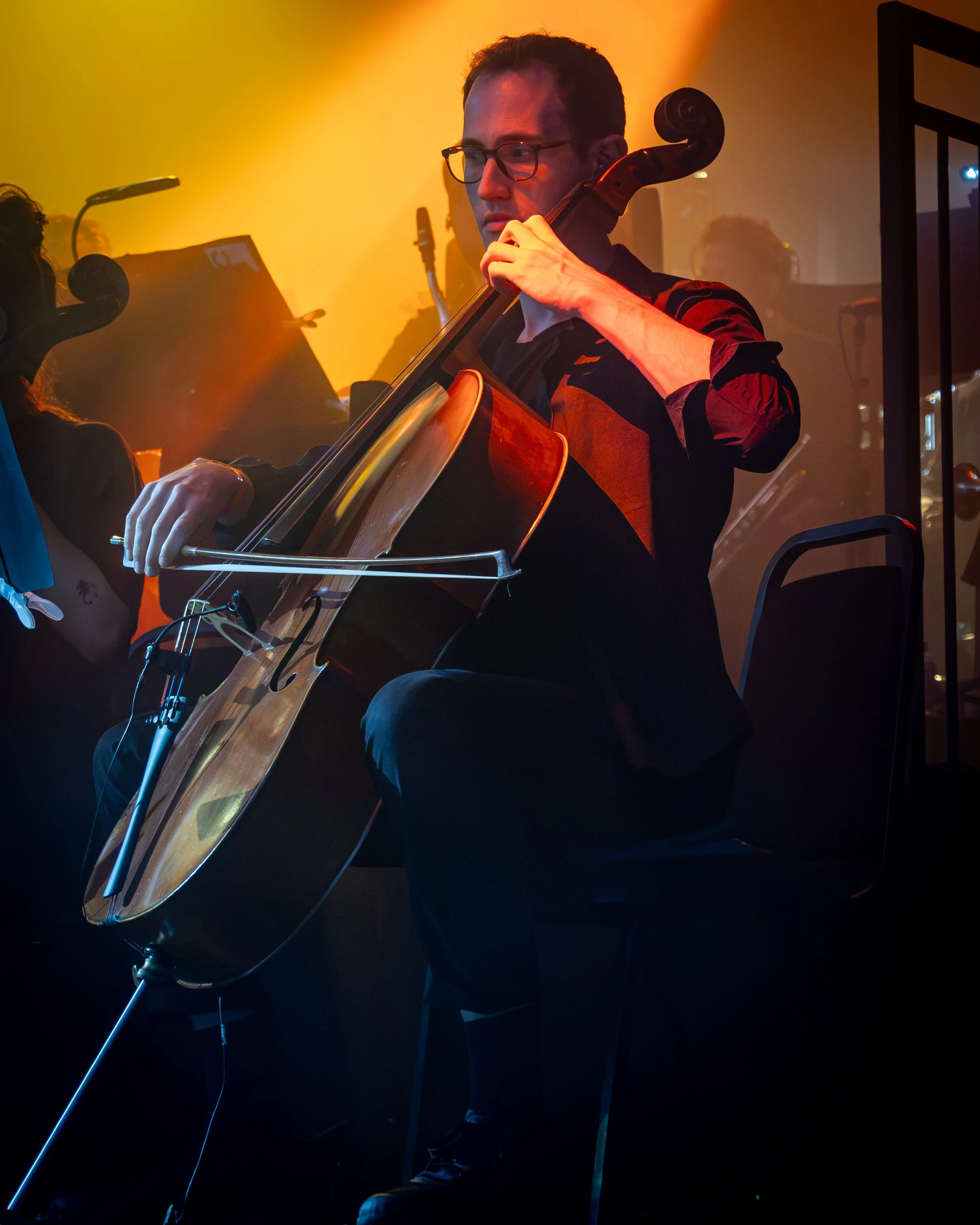A man wearing glasses and a black and red shirt playing a cello in a dimly lit setting with orange and blue lighting.
