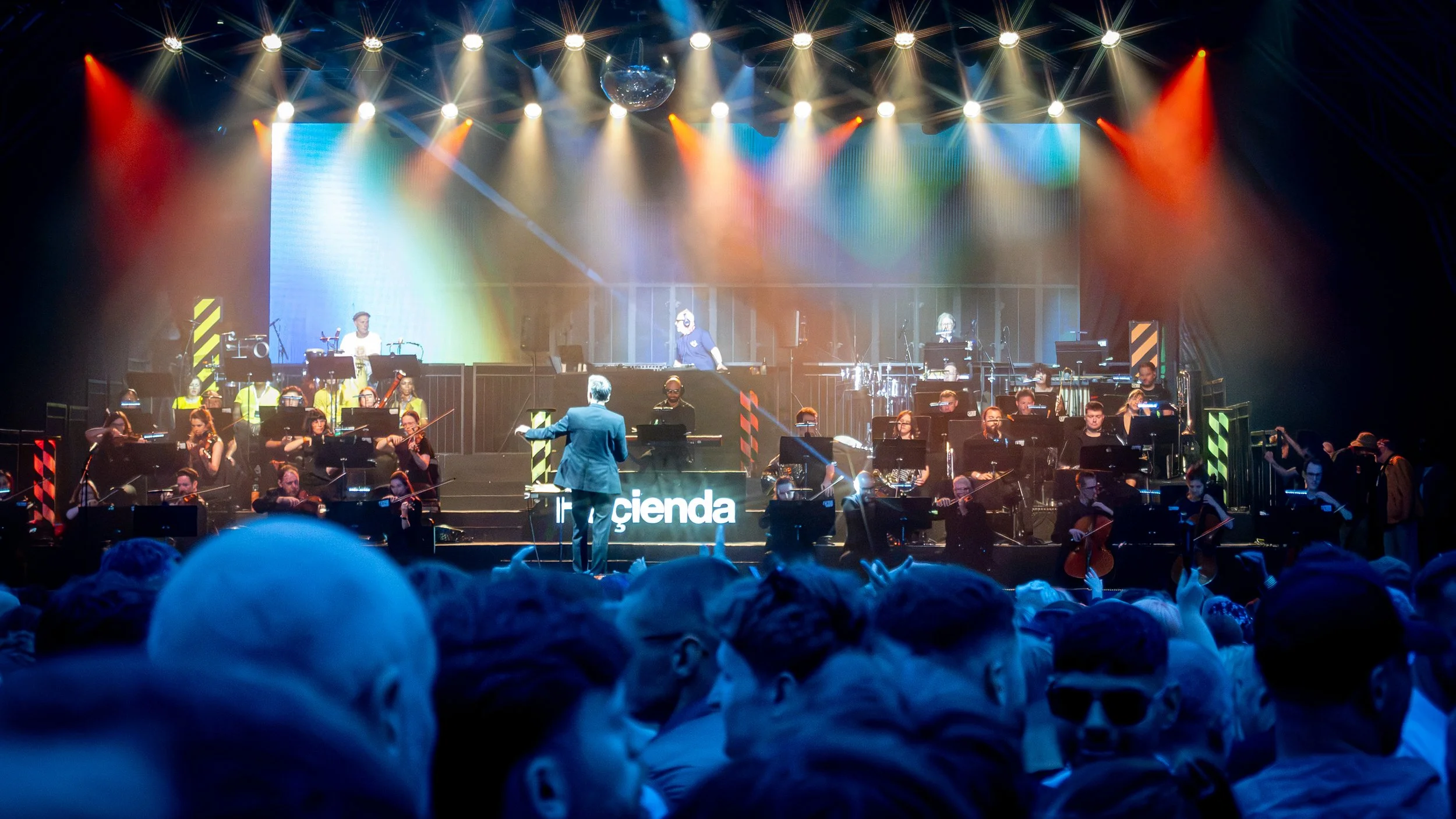 A large orchestra performing on stage, with a conductor in front, surrounded by colorful stage lighting and a digital screen in the background. Audience members are visible in the foreground.