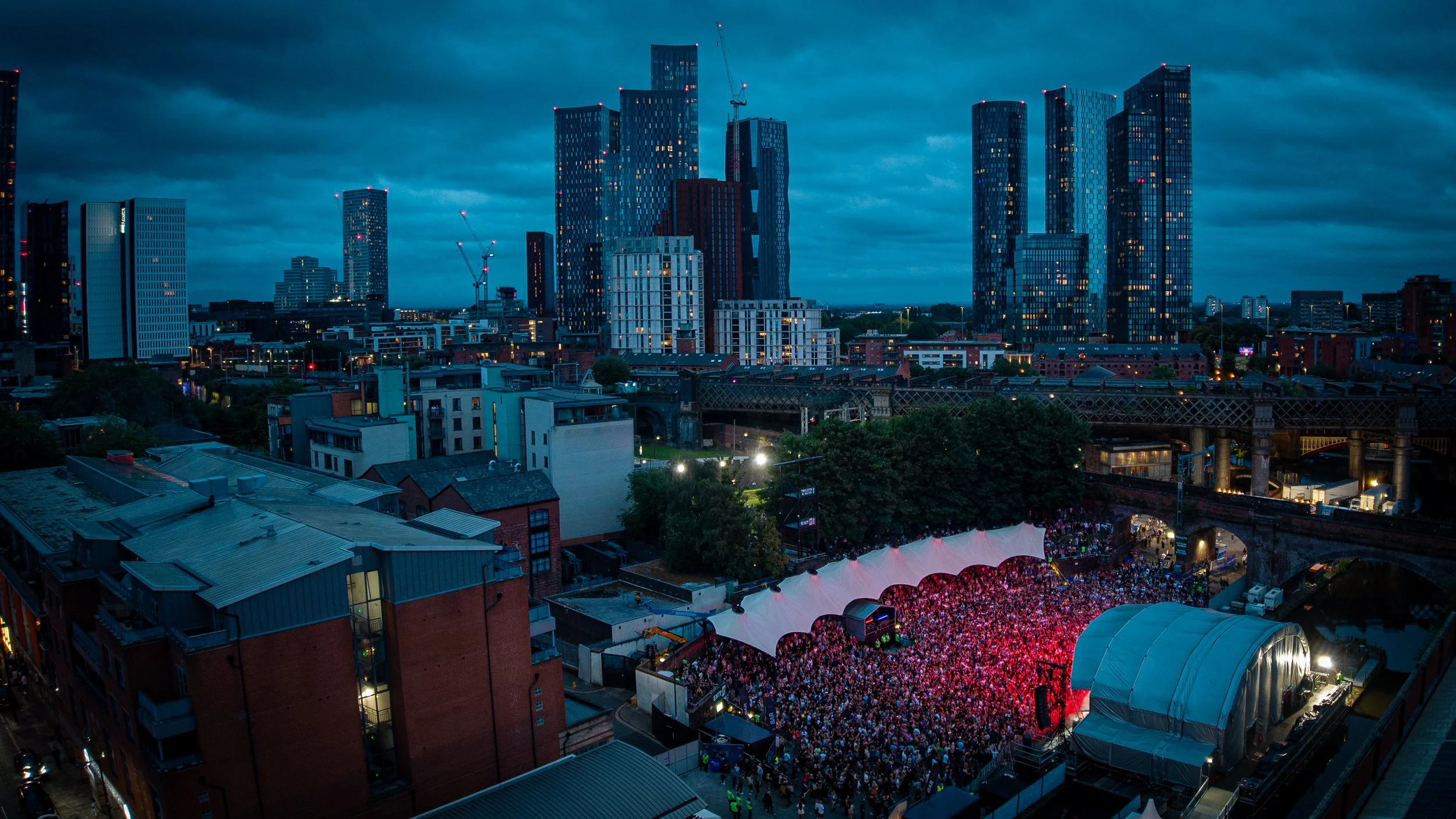 City skyline at dusk with tall modern buildings, construction cranes, and a large outdoor concert with a crowded audience beneath a tent.