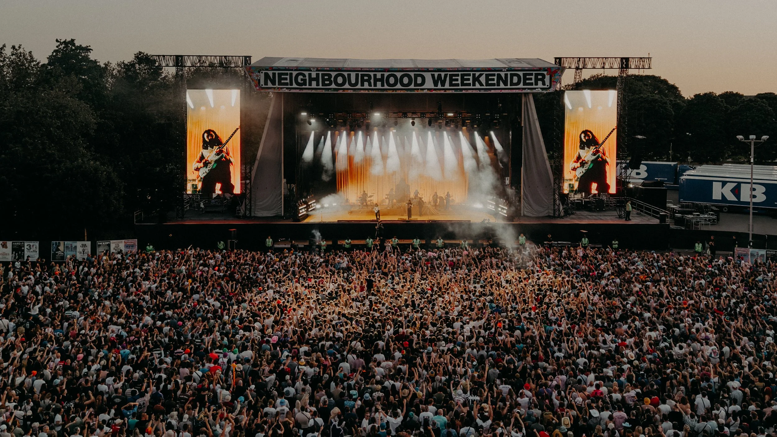 Large crowd attending an outdoor concert stage with a sign that reads 'Neighborhood Weekender,' with two large screens showing performers, smoke effects, and stage lighting.