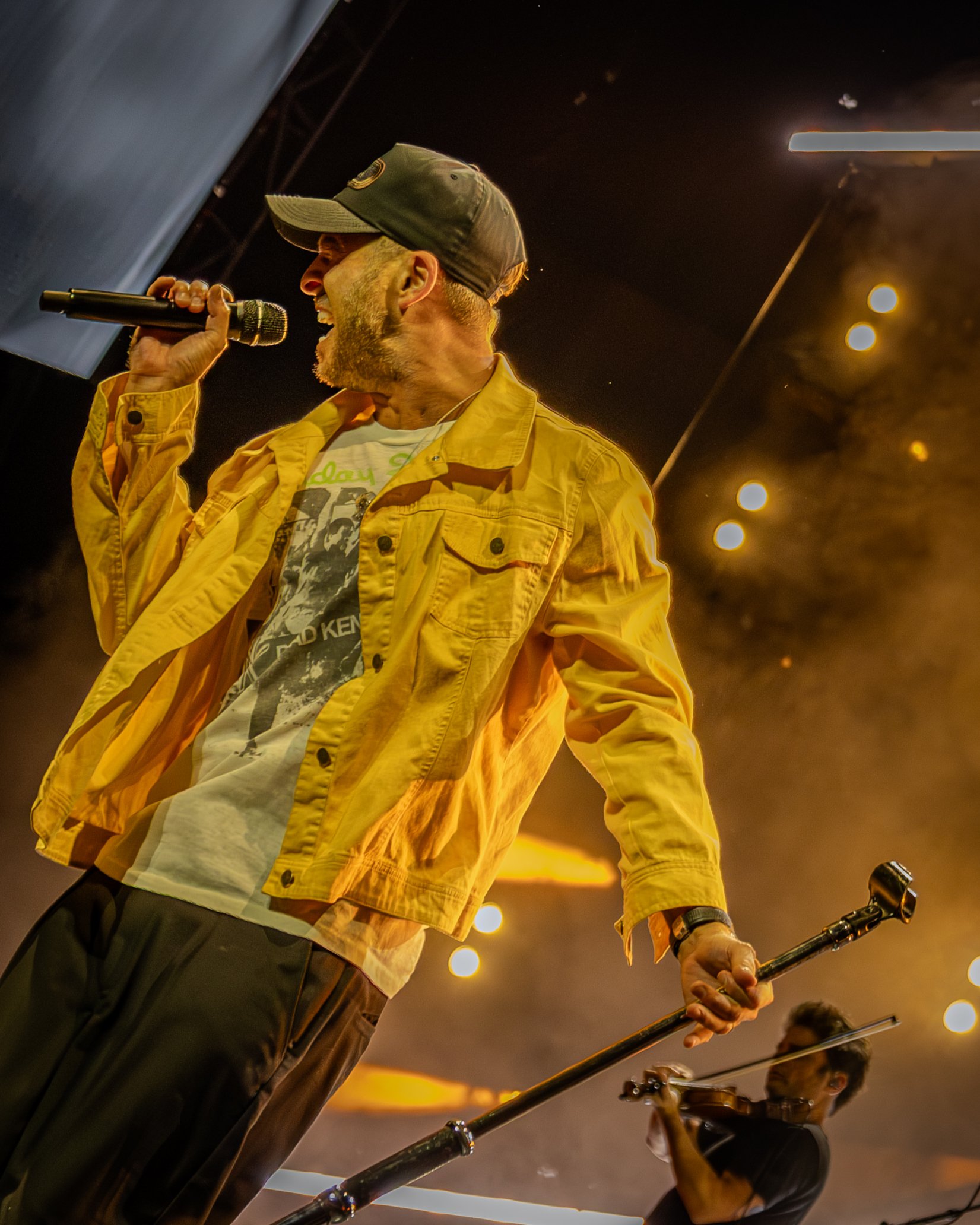 Man wearing a yellow jacket and cap singing into a microphone while playing a steel drum in an indoor setting with warm lighting.