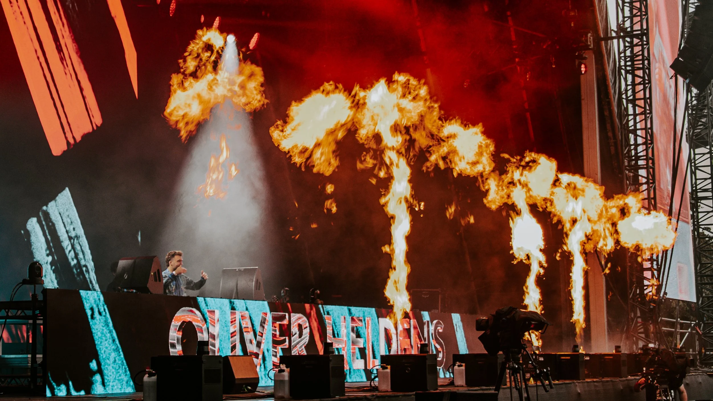 A concert stage with flames erupting from large flame projectors, a DJ or performer on stage, and a colorful LED screen display behind them.