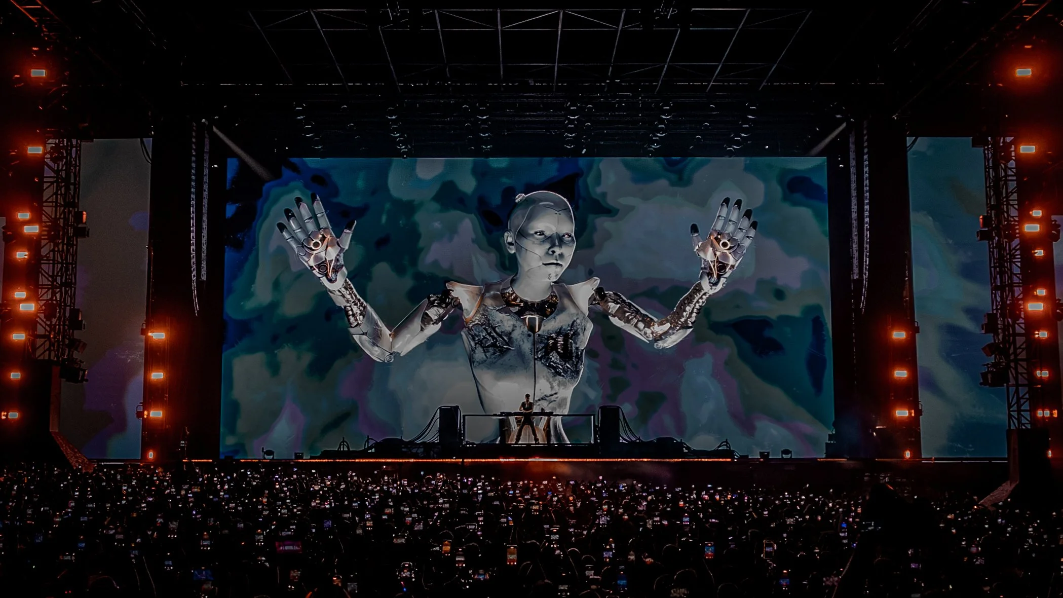 Large concert stage with an animated robot figure projected on the screen behind the DJ, surrounded by orange stage lights, with a crowd in front.