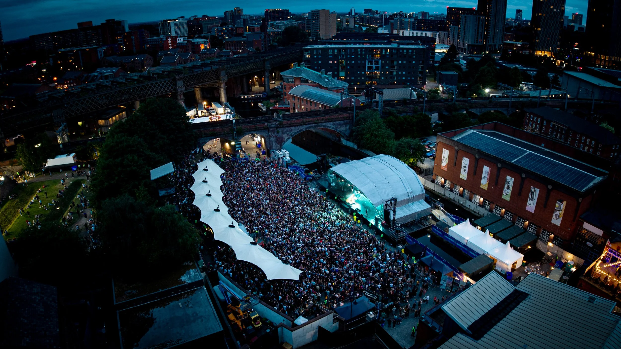 Aerial view of a city at night with a large crowd attending an outdoor concert, illuminated tents, and surrounding buildings.