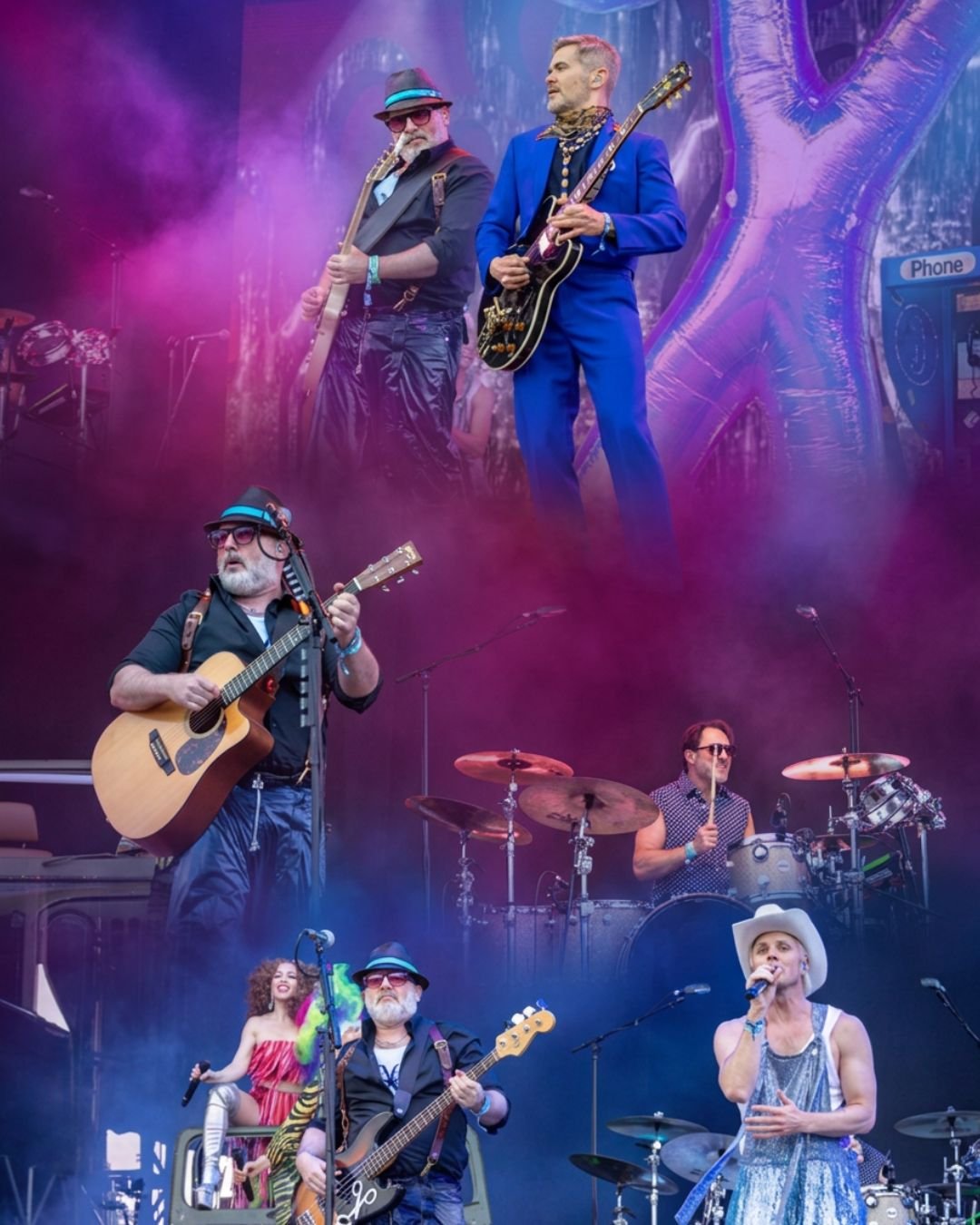 A colorful music band performing on stage with four members playing guitars, drums, and singing, surrounded by purple and pink stage lighting and fog effects.