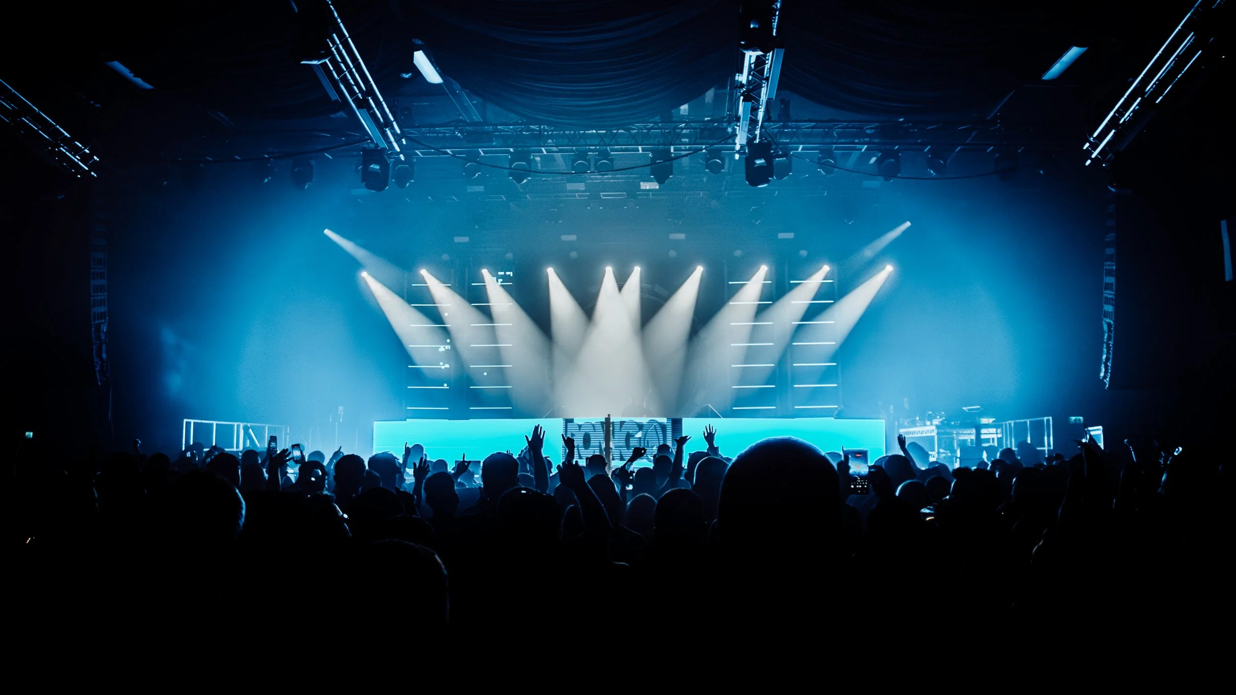 A concert scene with a large crowd facing a stage illuminated by blue lights and spotlights shining down. The audience has raised hands and some are taking photos or videos.