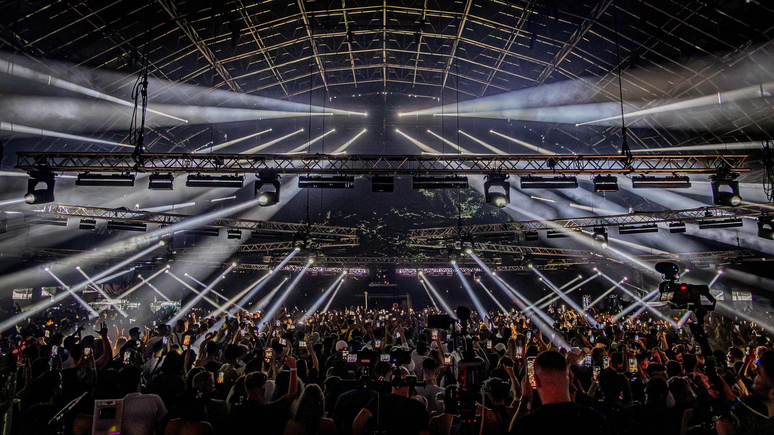 A large crowd at an indoor concert with bright white stage lights and fog effects overhead, as seen from the audience.