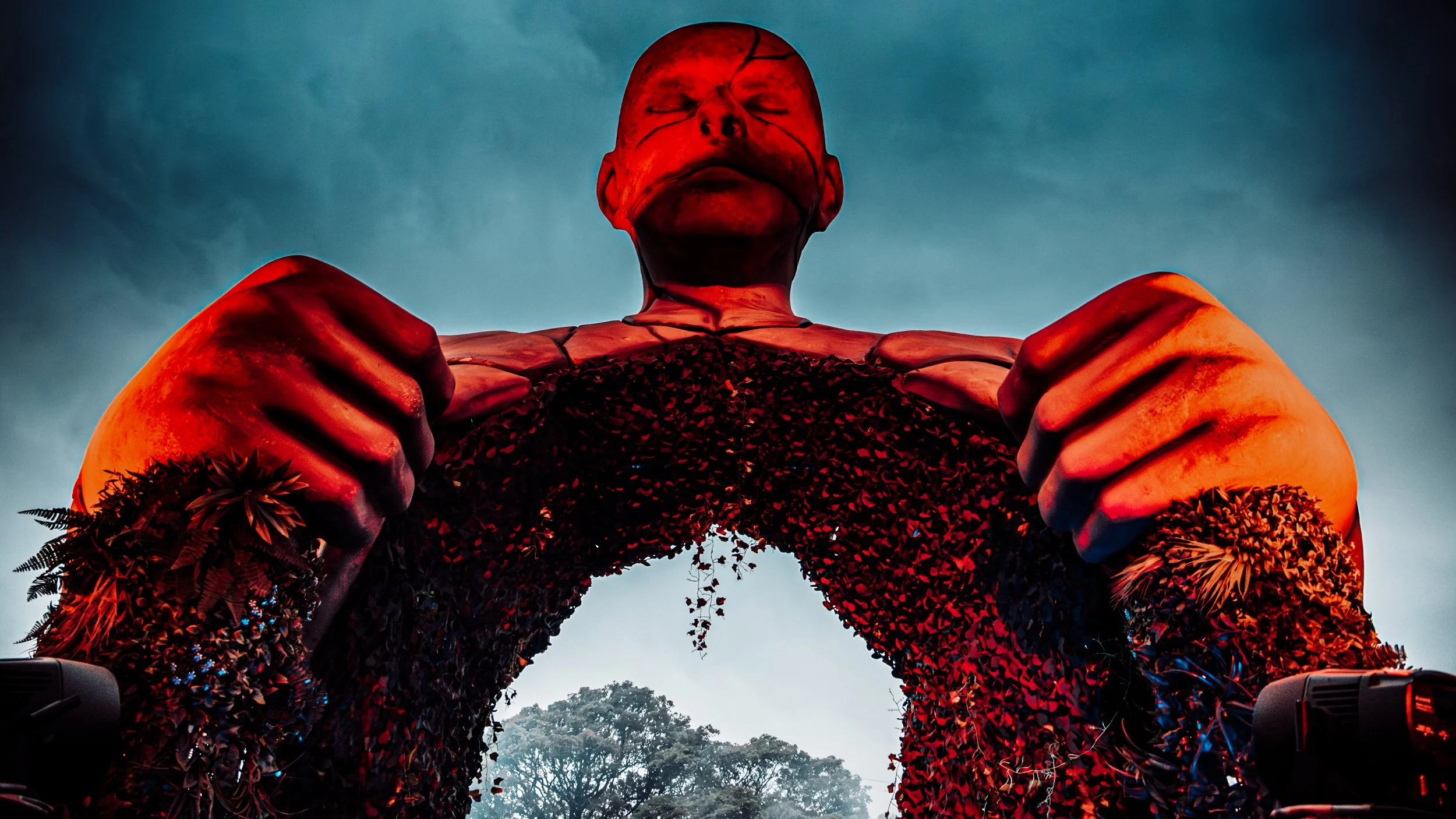 Large red humanoid sculpture of a person holding a circular object, with a cloudy sky and trees in the background.