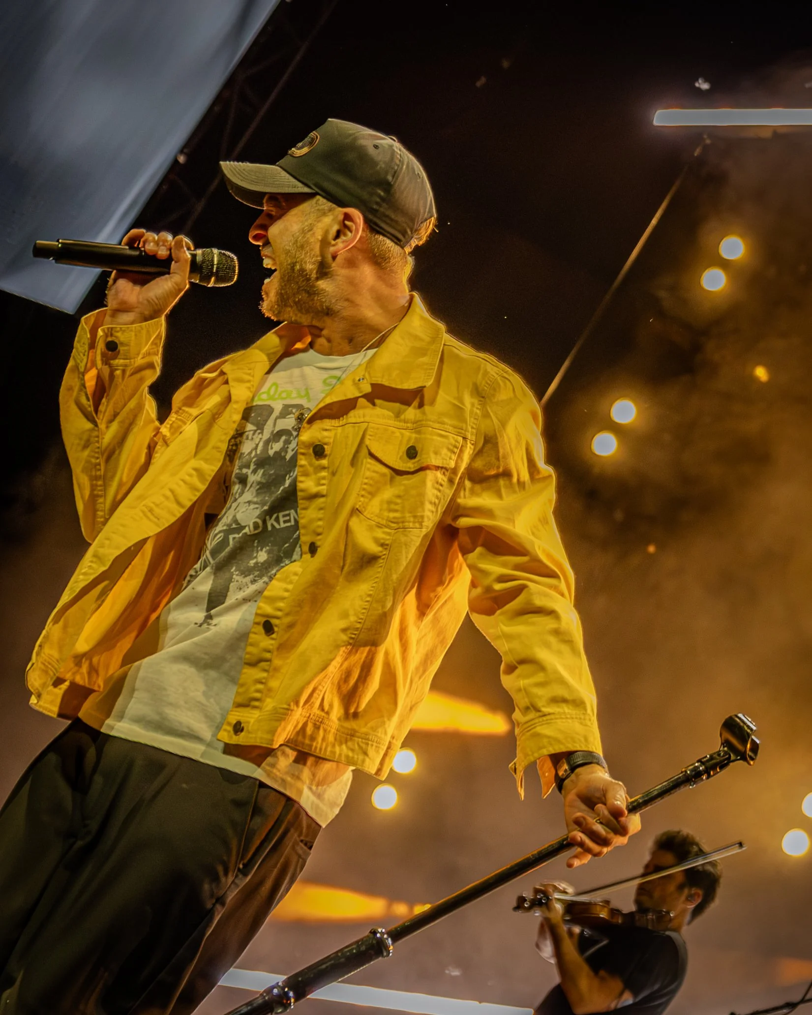 A man in a yellow jacket and cap playing a steel drum, with another person in the background playing a string instrument, at sunset with warm lighting and bokeh effects.