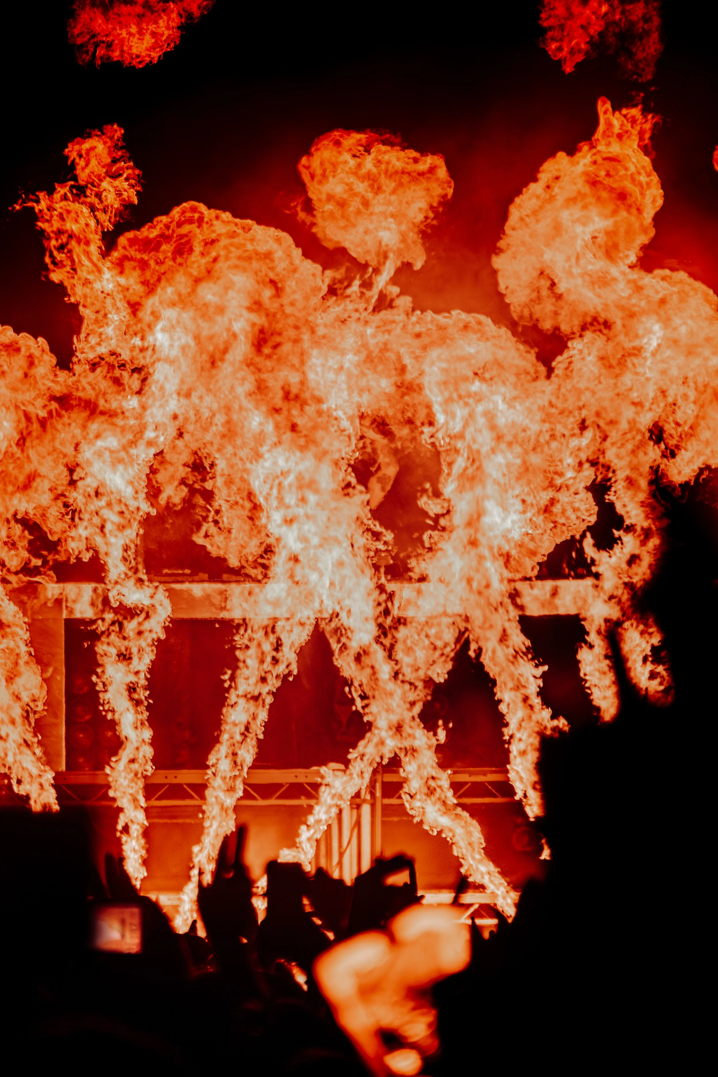 Large flames and fireballs erupt in the night sky, with silhouettes of people in the foreground watching the fiery display.