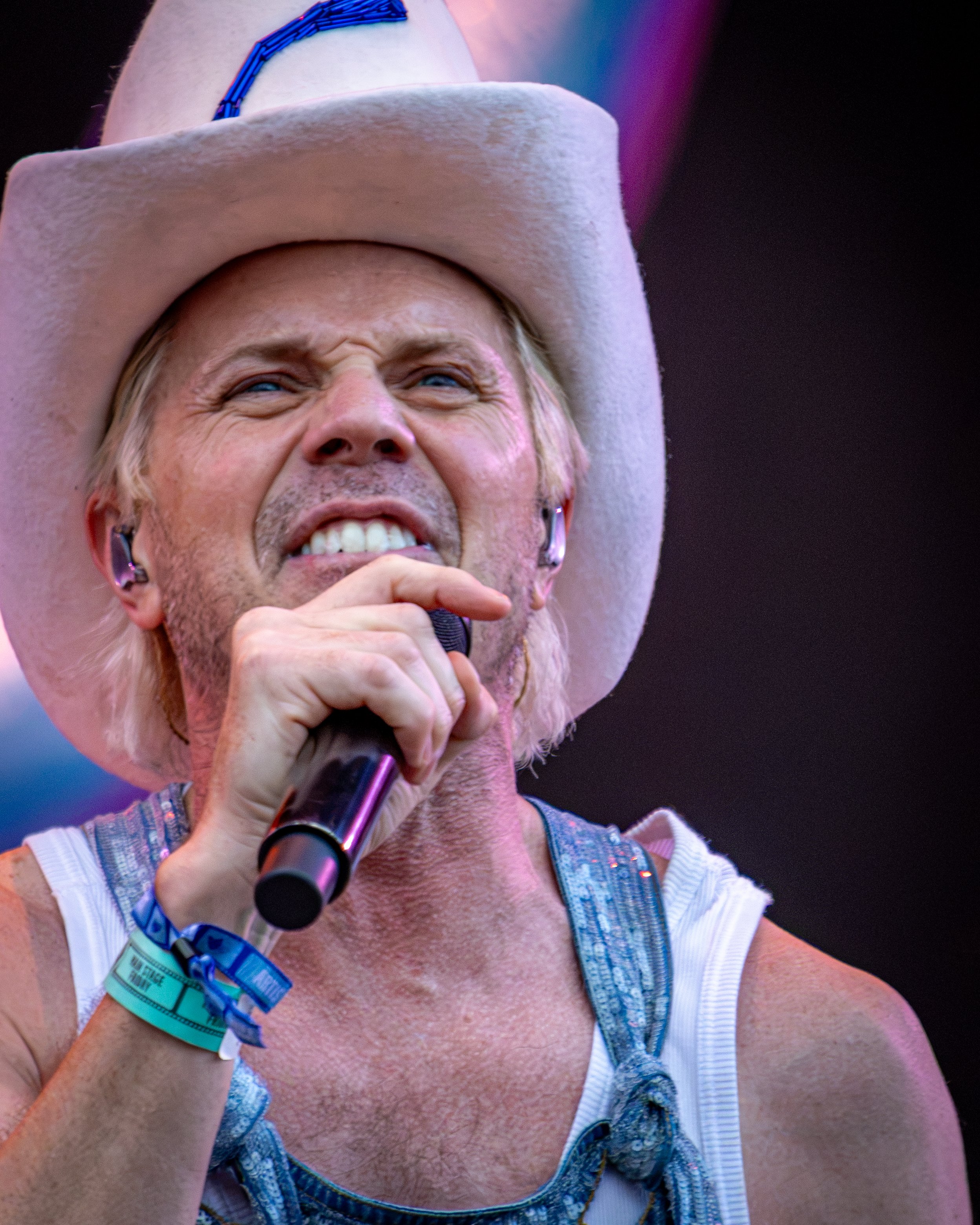 Close-up of a smiling person wearing a cowboy hat, denim overalls, holding a microphone, with backstage or stage lighting in the background.