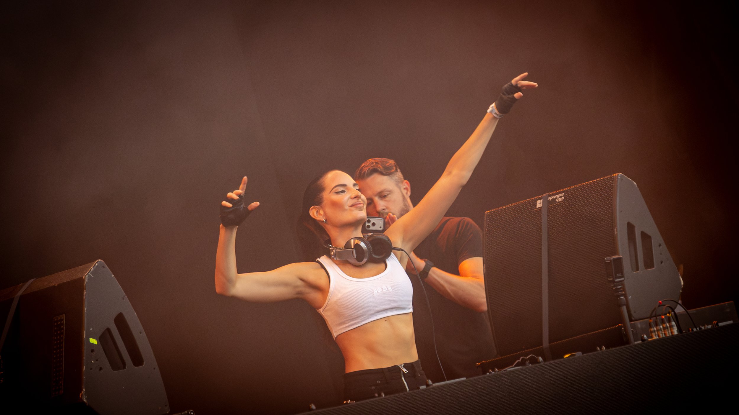 A female DJ with dark hair, wearing a white crop top and headphones around her neck, raising her arms with a confident expression while standing behind DJ equipment. A male assistant or fellow DJ stands behind her in a dark environment with stage lig