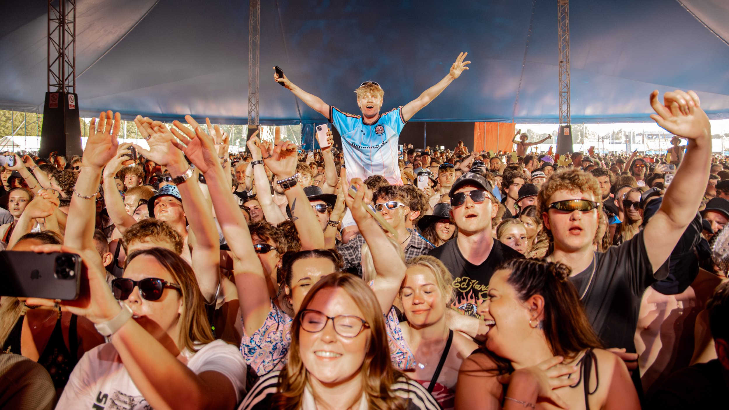 Crowd of people enjoying a concert, with someone sitting on shoulders of others, taking selfies and wearing sunglasses inside a large tent.