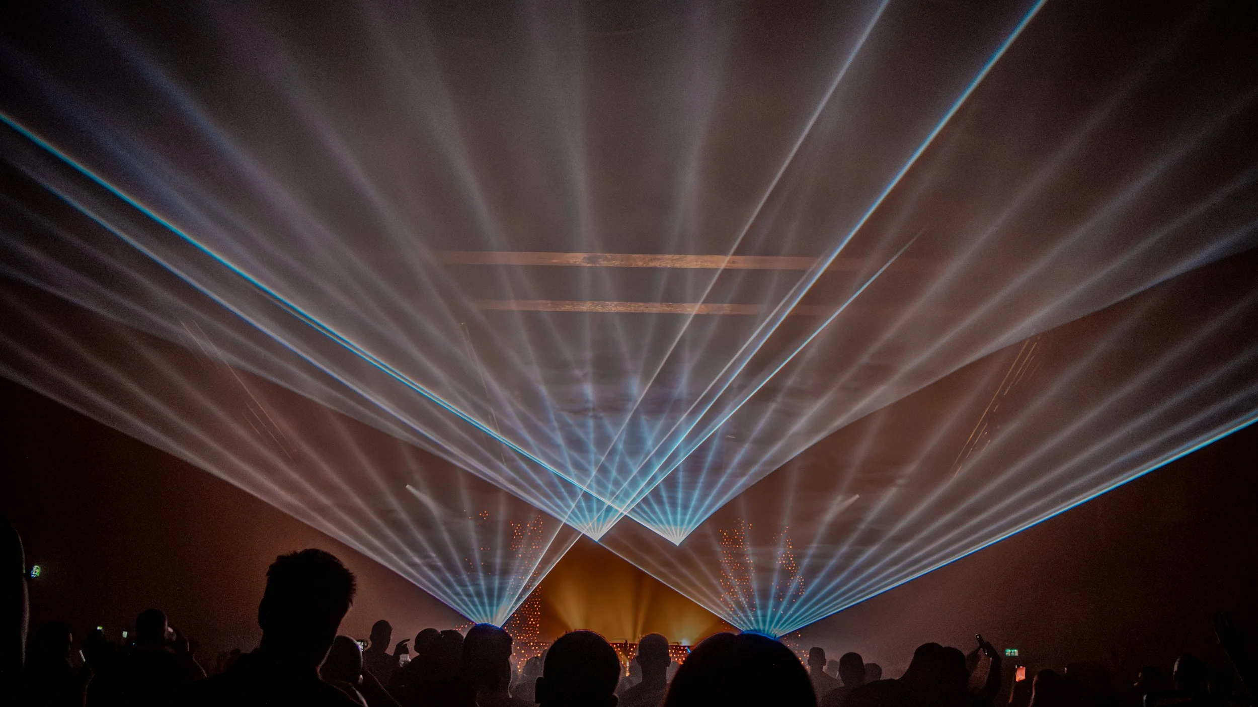 A crowd of people watching a laser light show with multiple beams of blue and white light radiating outward in a dark environment.