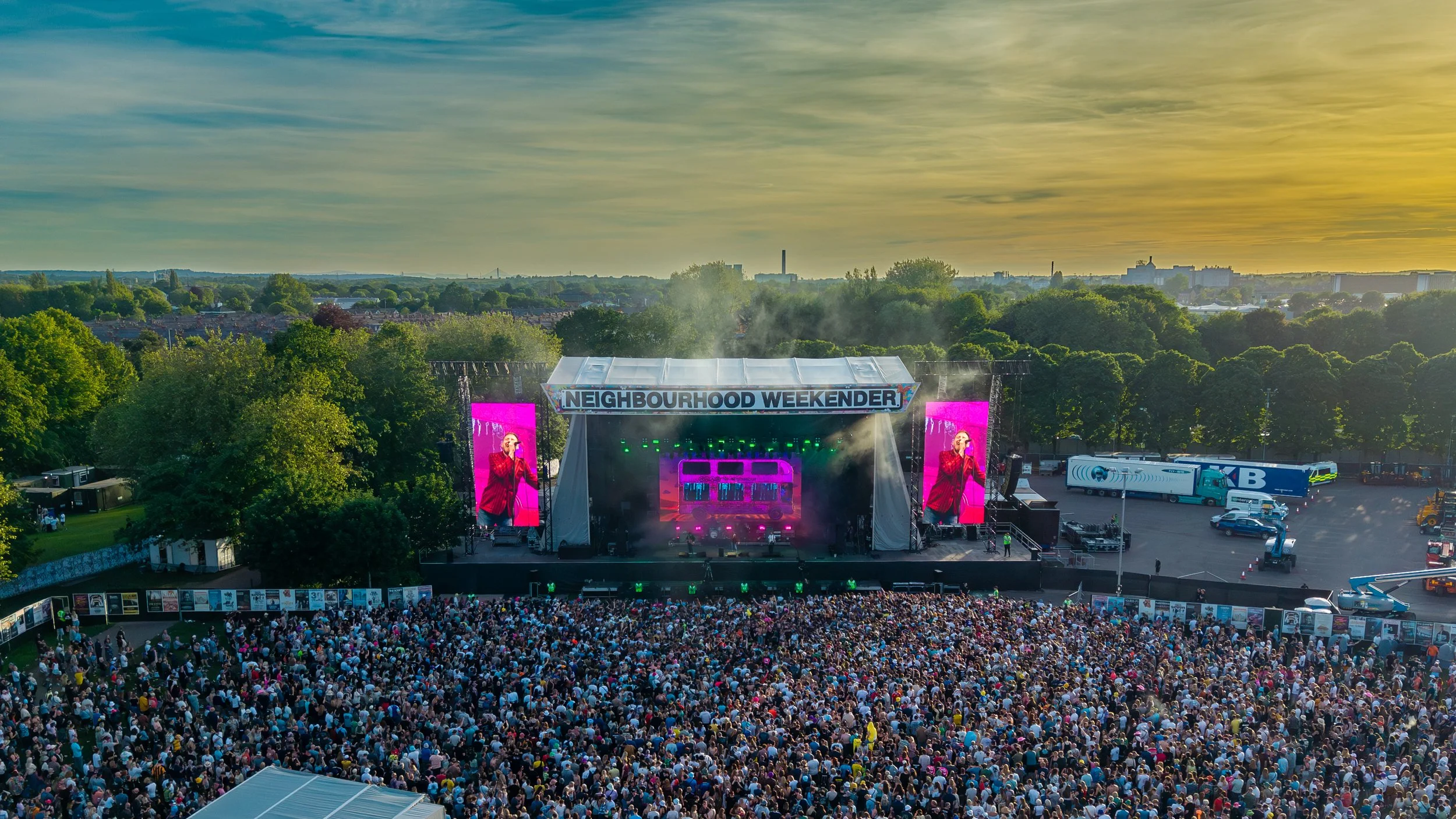 Outdoor concert stage with a large crowd watching performers, trees surrounding the area, and a cityscape in the background at sunset. The stage has a sign reading 'NEIGHBOURHOOD WEEKENDER' and large screens displaying performers.