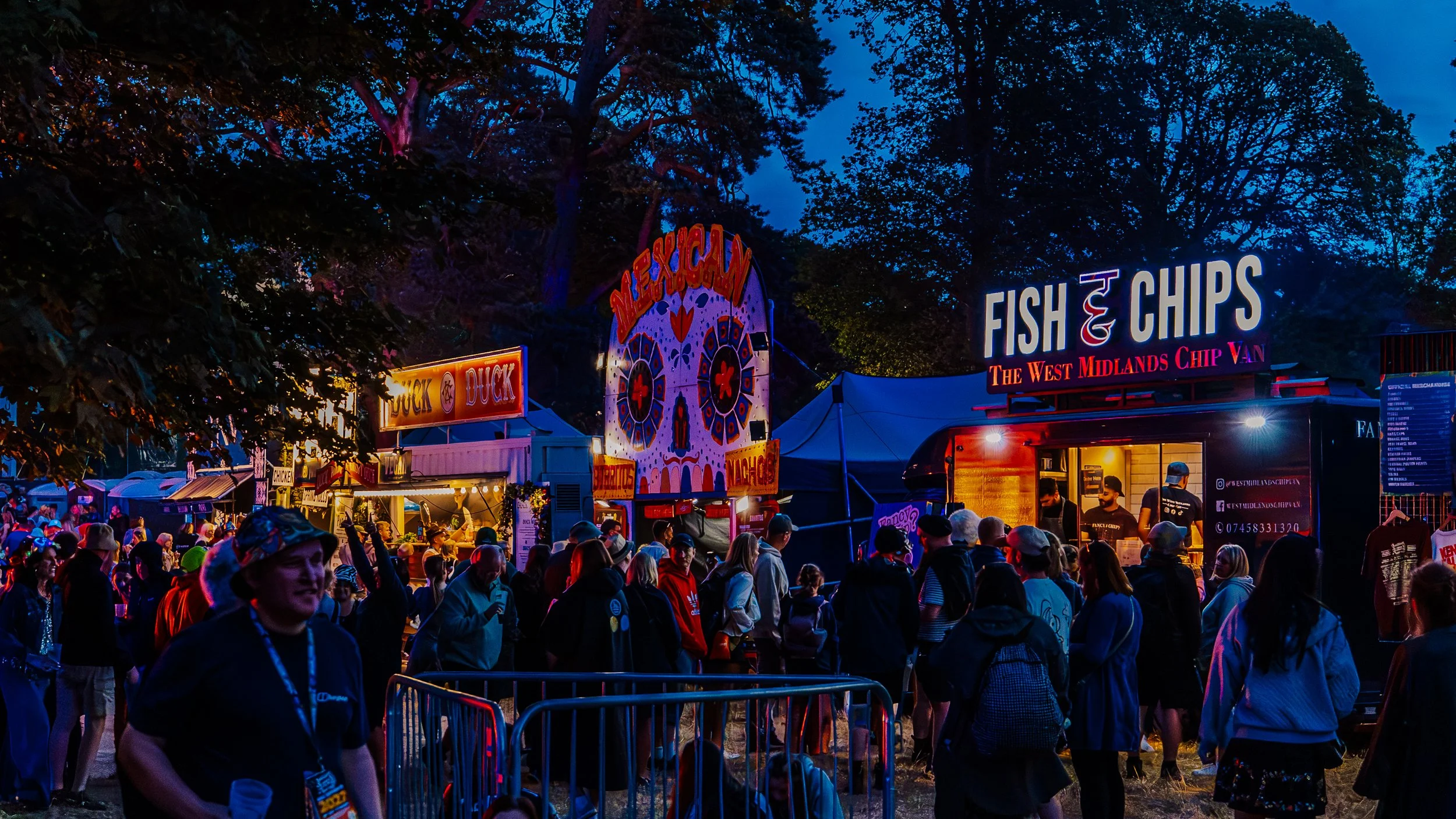 Night scene at a fair with food stalls, bright signs, and a large crowd of people, including a food truck selling fish and chips, surrounded by trees.