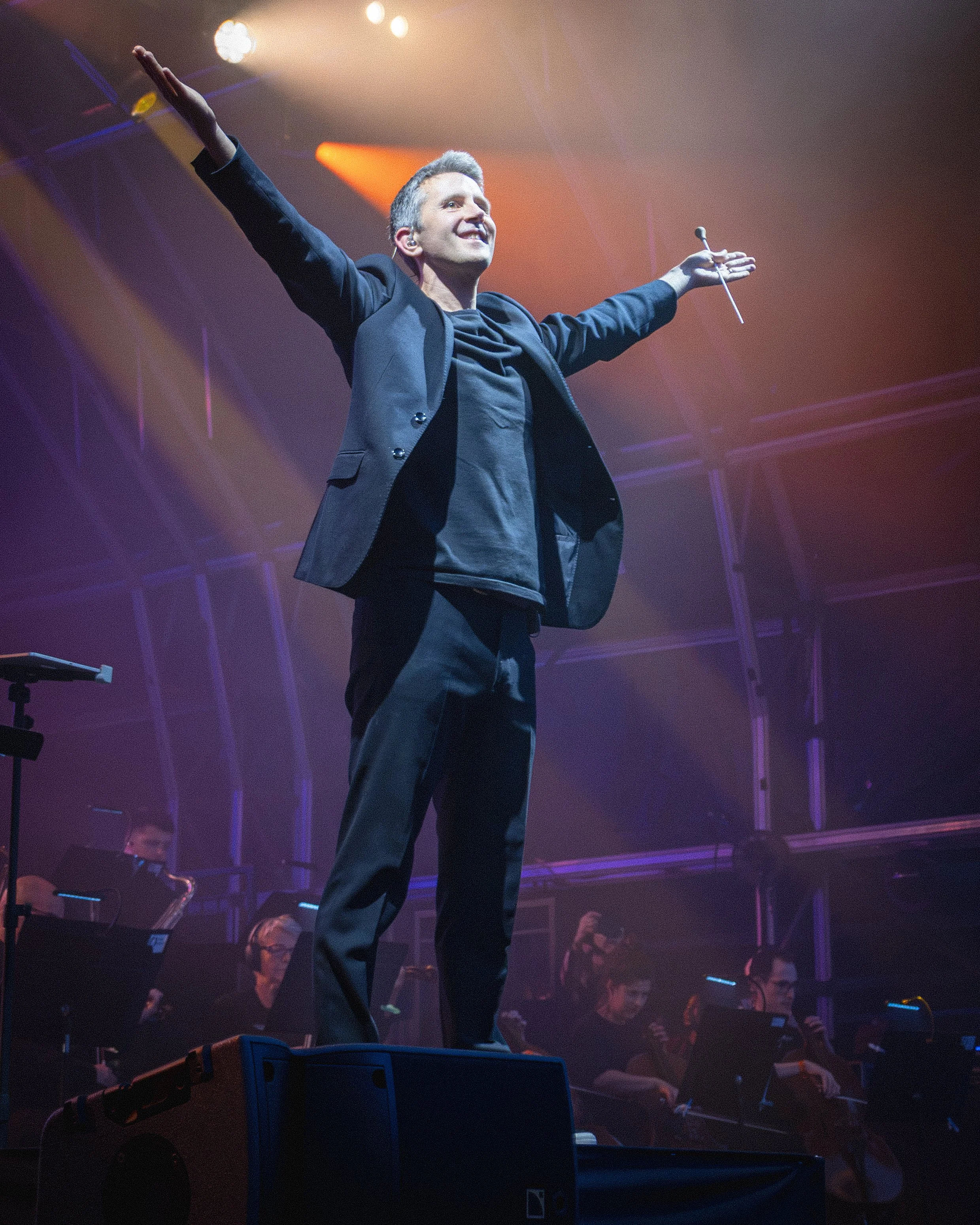 A man with gray hair, wearing a black jacket and dark pants, stands on stage with his arms outstretched, holding a conductor's baton in his right hand. He is smiling and looking upward, with stage lights creating a colorful backdrop.