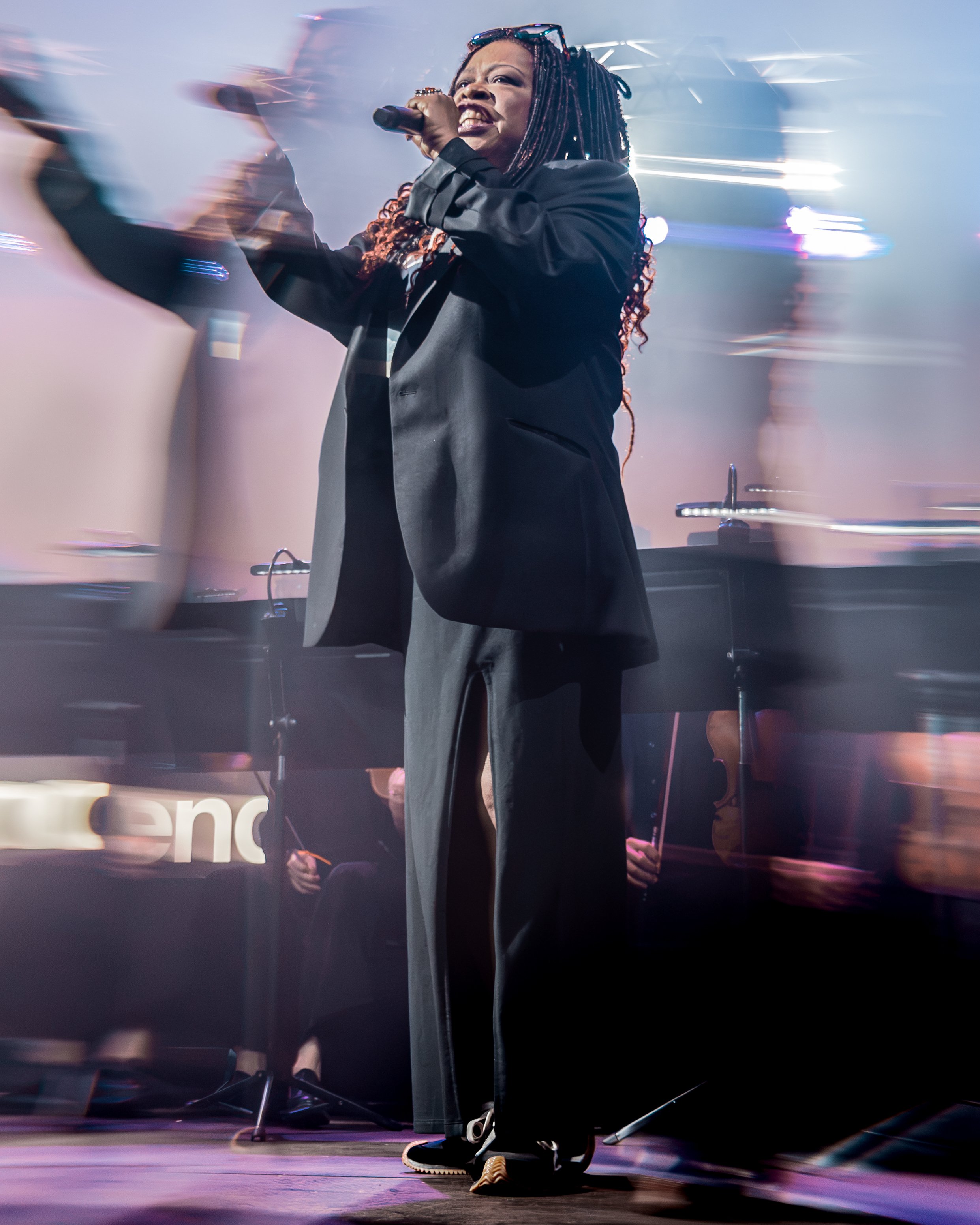 A woman with braided hair singing passionately into a microphone on stage with vibrant lighting and smoke effects.