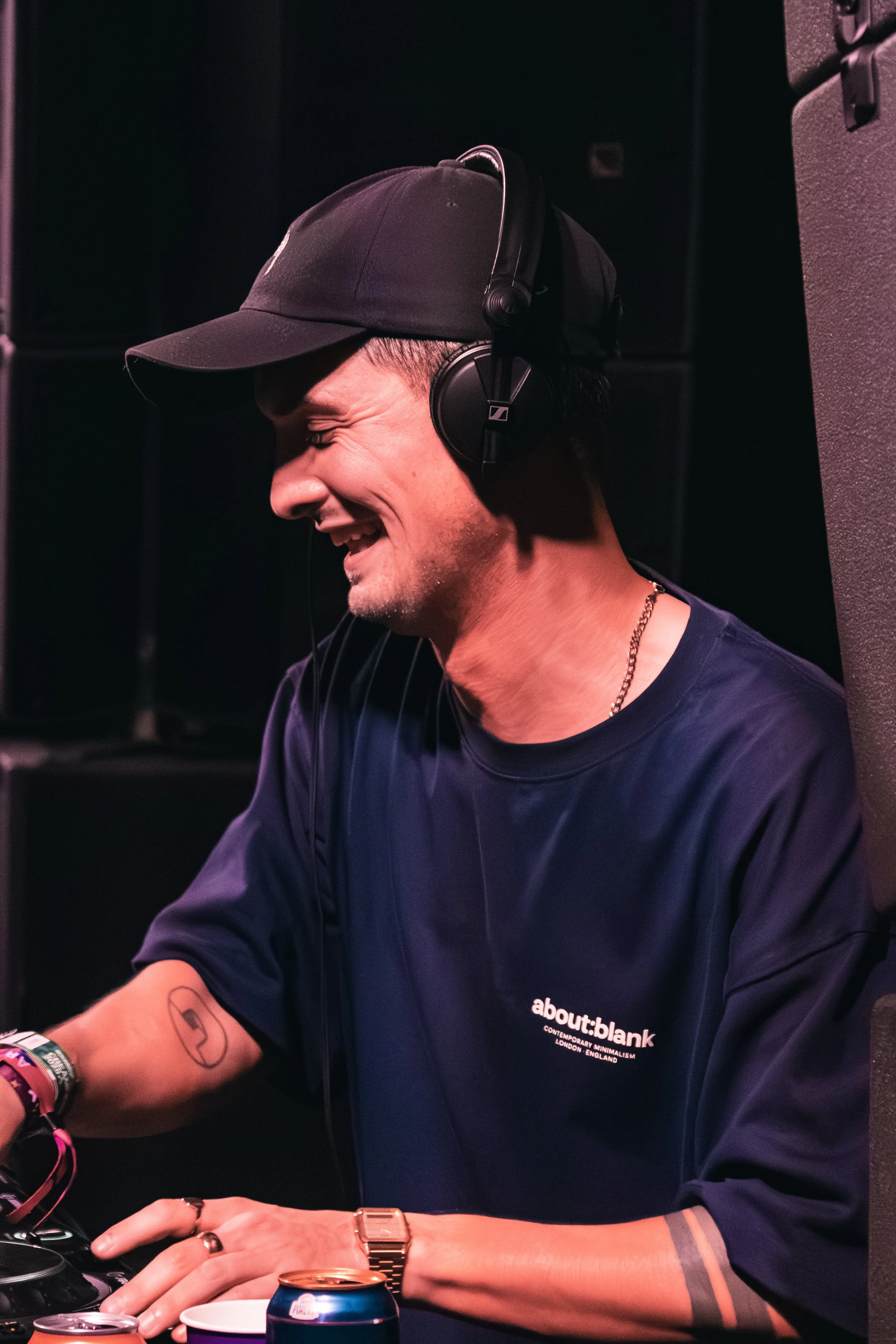 A person wearing a black baseball cap, black headphones, a black t-shirt, and jewelry, smiling with eyes closed, sitting at a table with drinks, and a tattoo on their forearm.