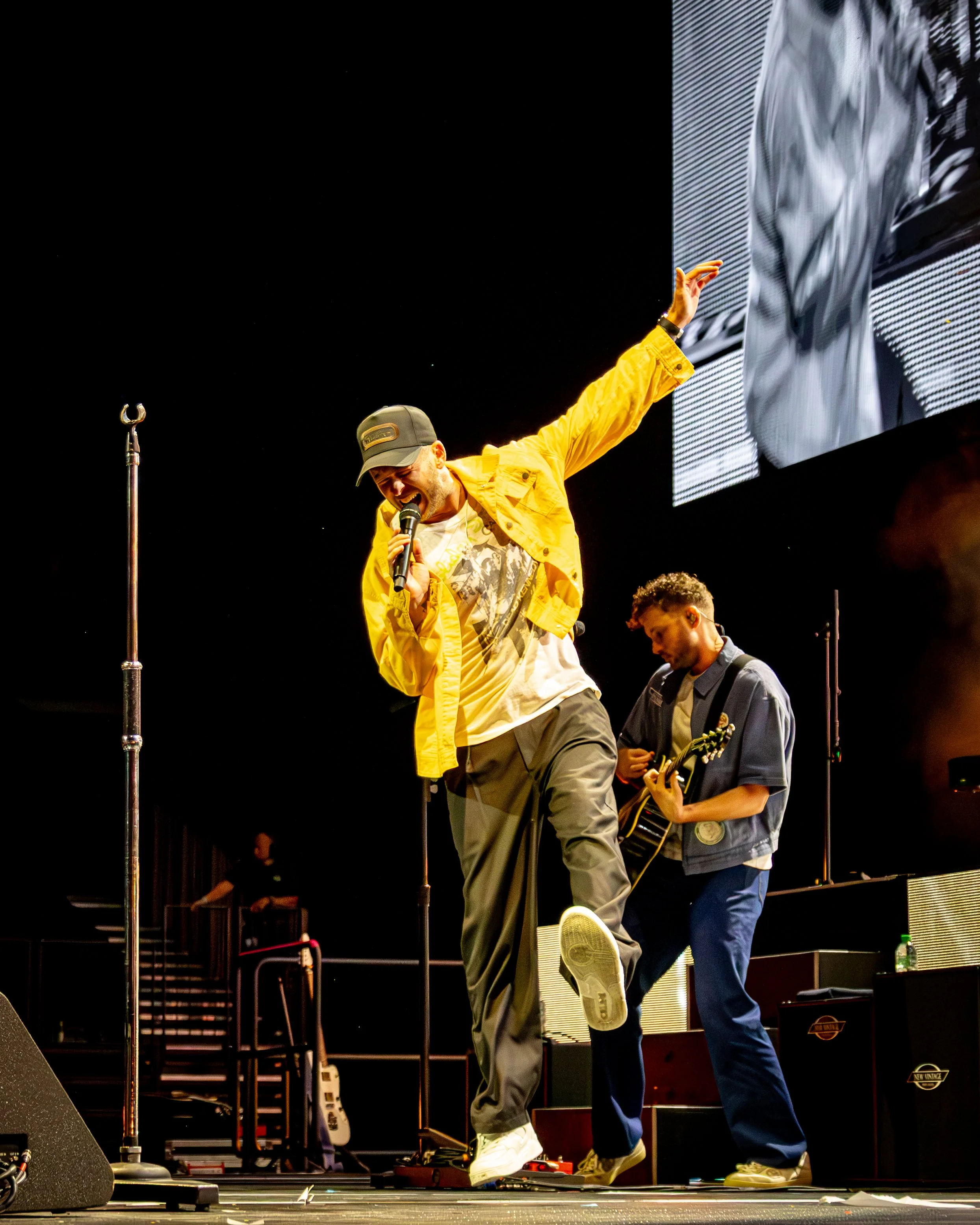 Performer in a yellow jacket singing into a microphone on stage, with a large screen in the background and musicians playing instruments nearby.